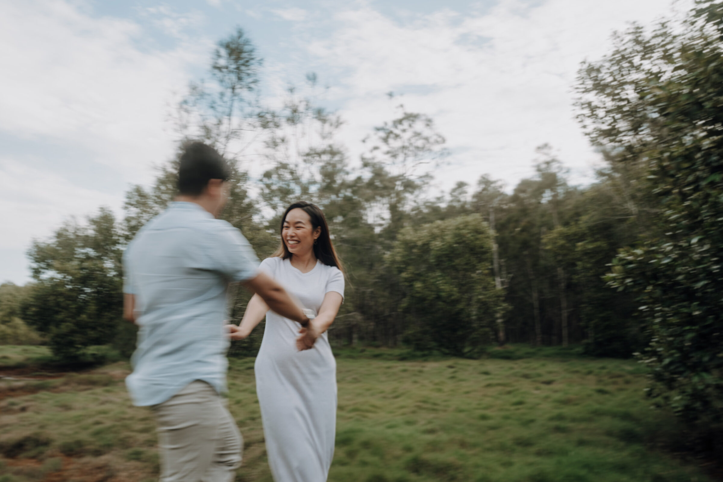 A couple in casual attire playfully spins in a grassy field, surrounded by trees under a cloudy sky.