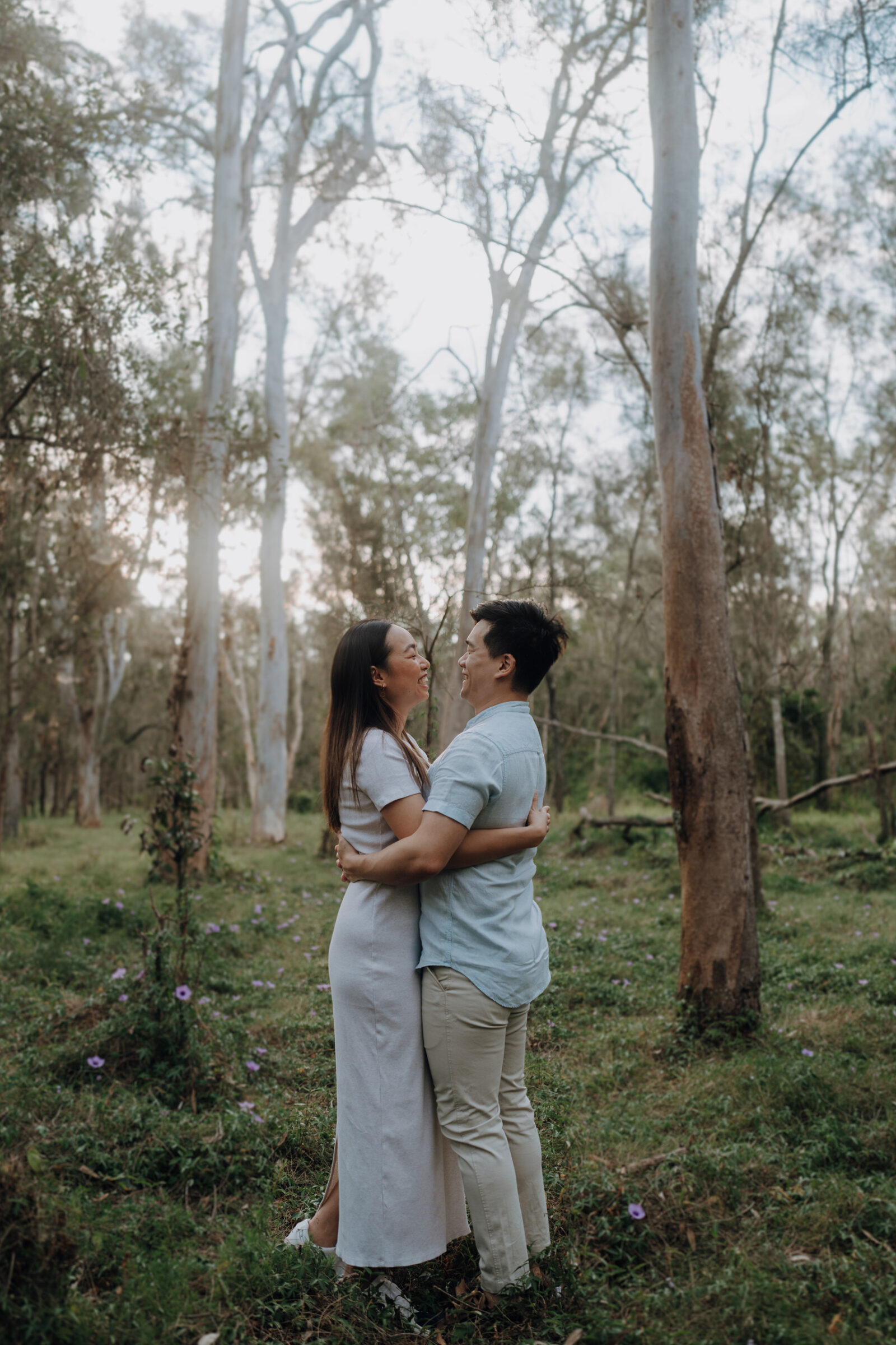 A couple embraces and laughs in a forested area, surrounded by tall trees and wildflowers.