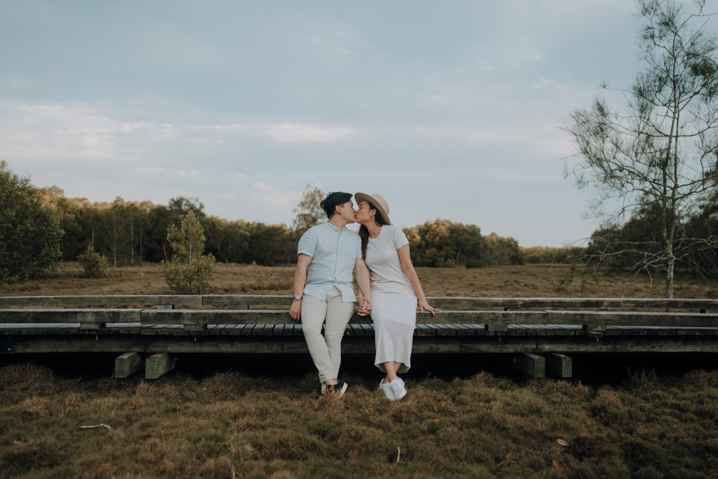 A couple sits on a wooden bridge in a grassy field, kissing under a cloudy sky. The woman wears a white dress and hat; the man wears a light blue shirt and beige pants.