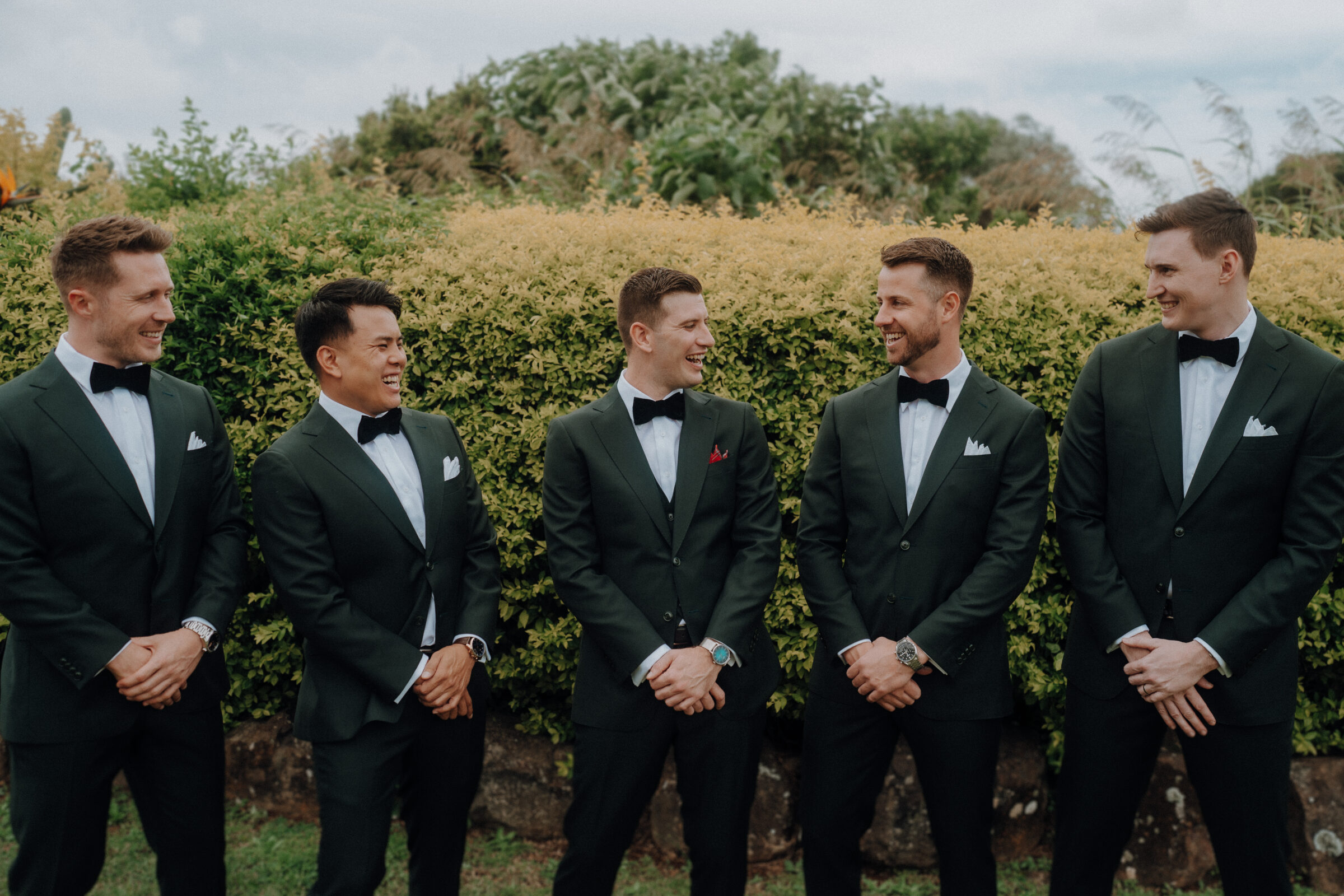 Five men in matching dark suits and bow ties stand outdoors in front of a hedge, smiling and conversing.