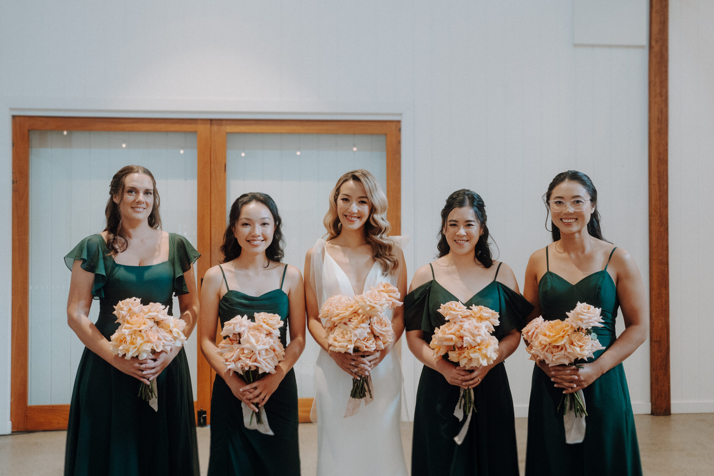 A bride in a white dress stands with four bridesmaids in matching green dresses, all holding bouquets of peach-colored flowers.