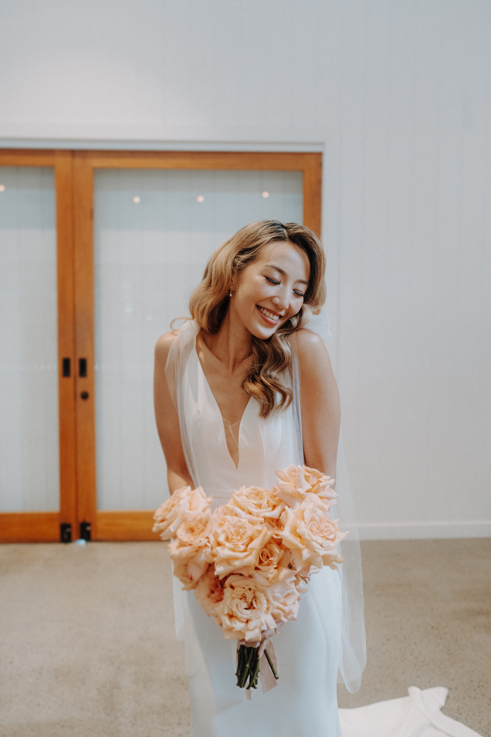 Bride in a white dress smiles while holding a bouquet of peach roses, standing indoors with wooden double doors in the background.