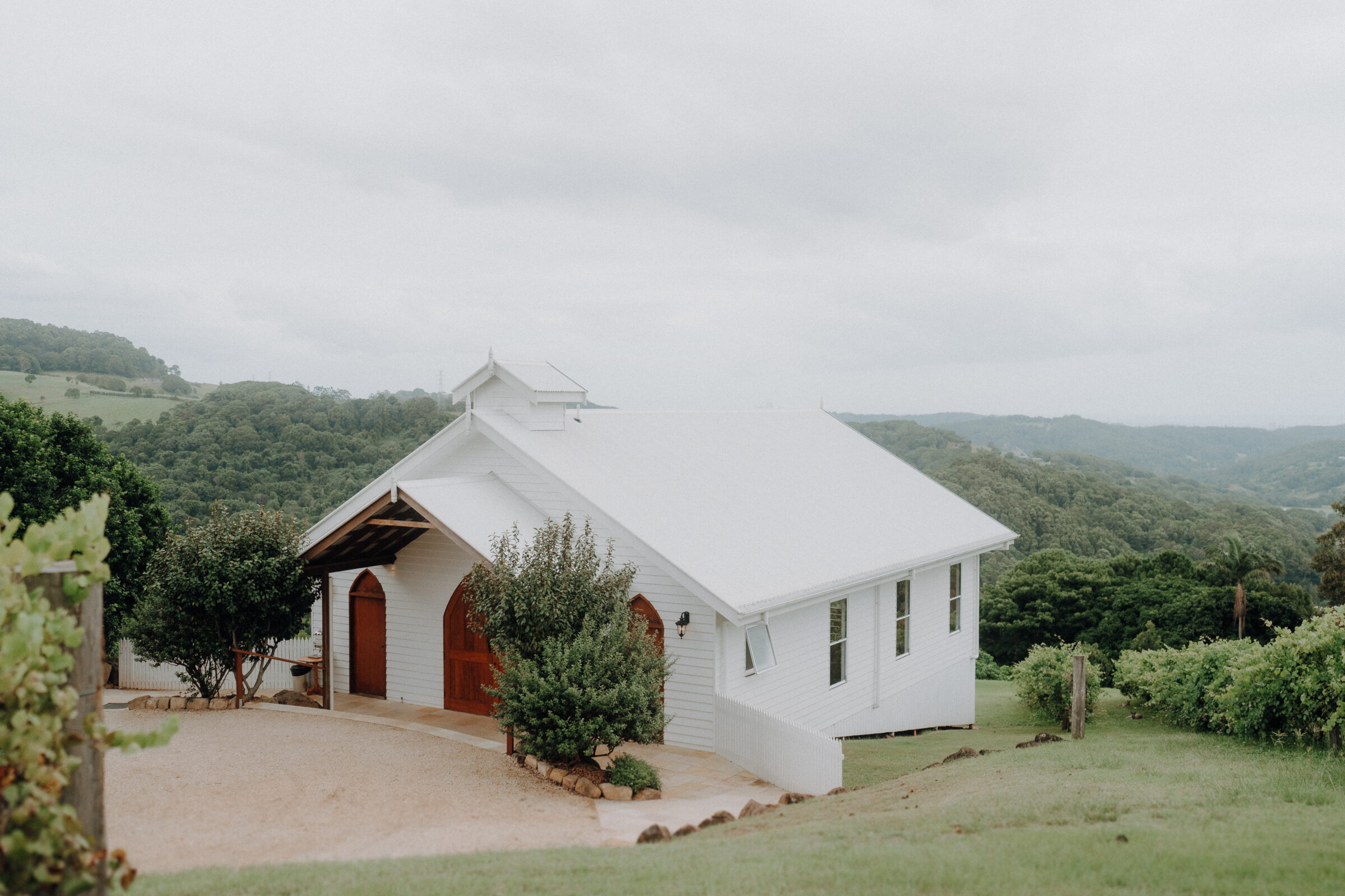 A small white chapel with a red door is surrounded by greenery and hills under a cloudy sky.