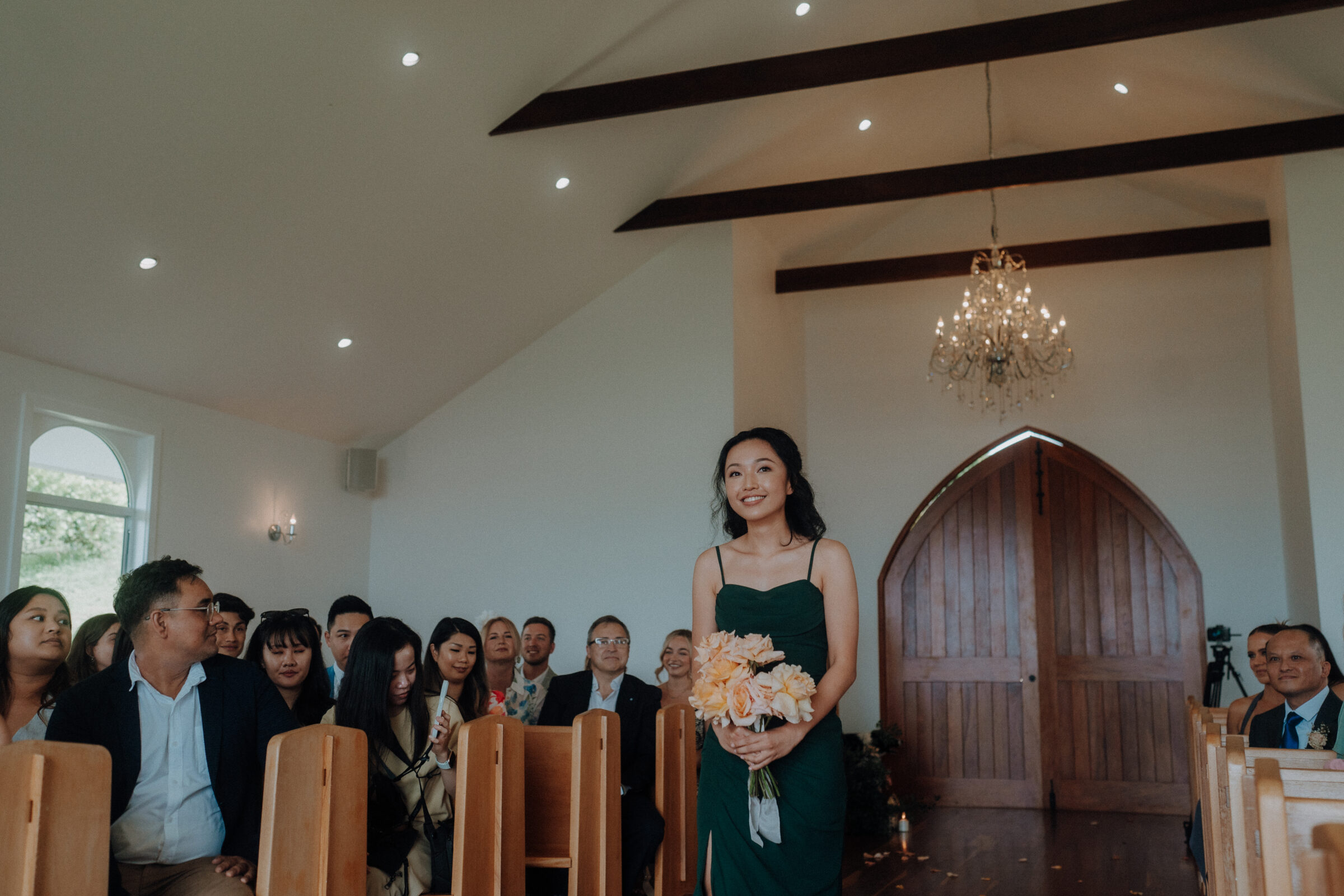 A woman in a green dress walks down the aisle of a church holding a bouquet. Guests are seated on wooden pews, and a chandelier hangs from the ceiling.