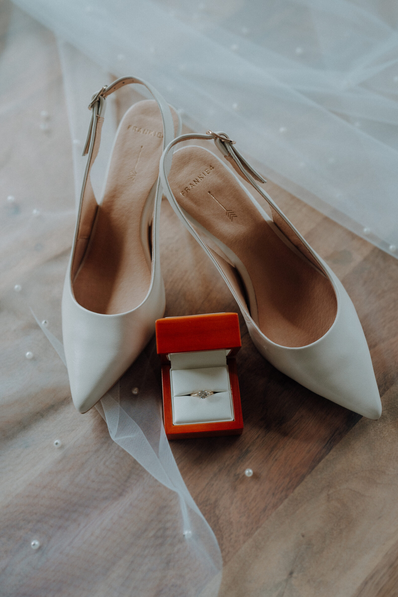 White high heels and a red ring box with a diamond ring placed on a sheer fabric decorated with small pearls.