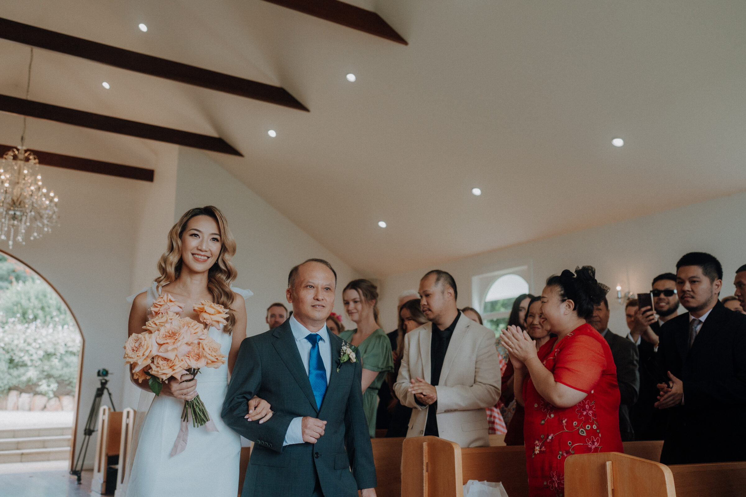 A bride in a white dress walks down the aisle with a man in a suit, surrounded by seated and standing guests in a chapel.