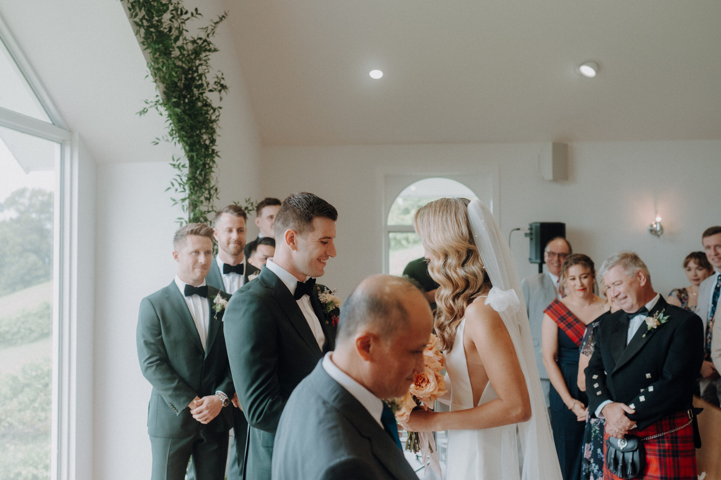 A couple stands at the altar during a wedding ceremony. The groom and groomsmen are in black suits, and the bride wears a white dress. Guests watch in a well-lit room with greenery decor.