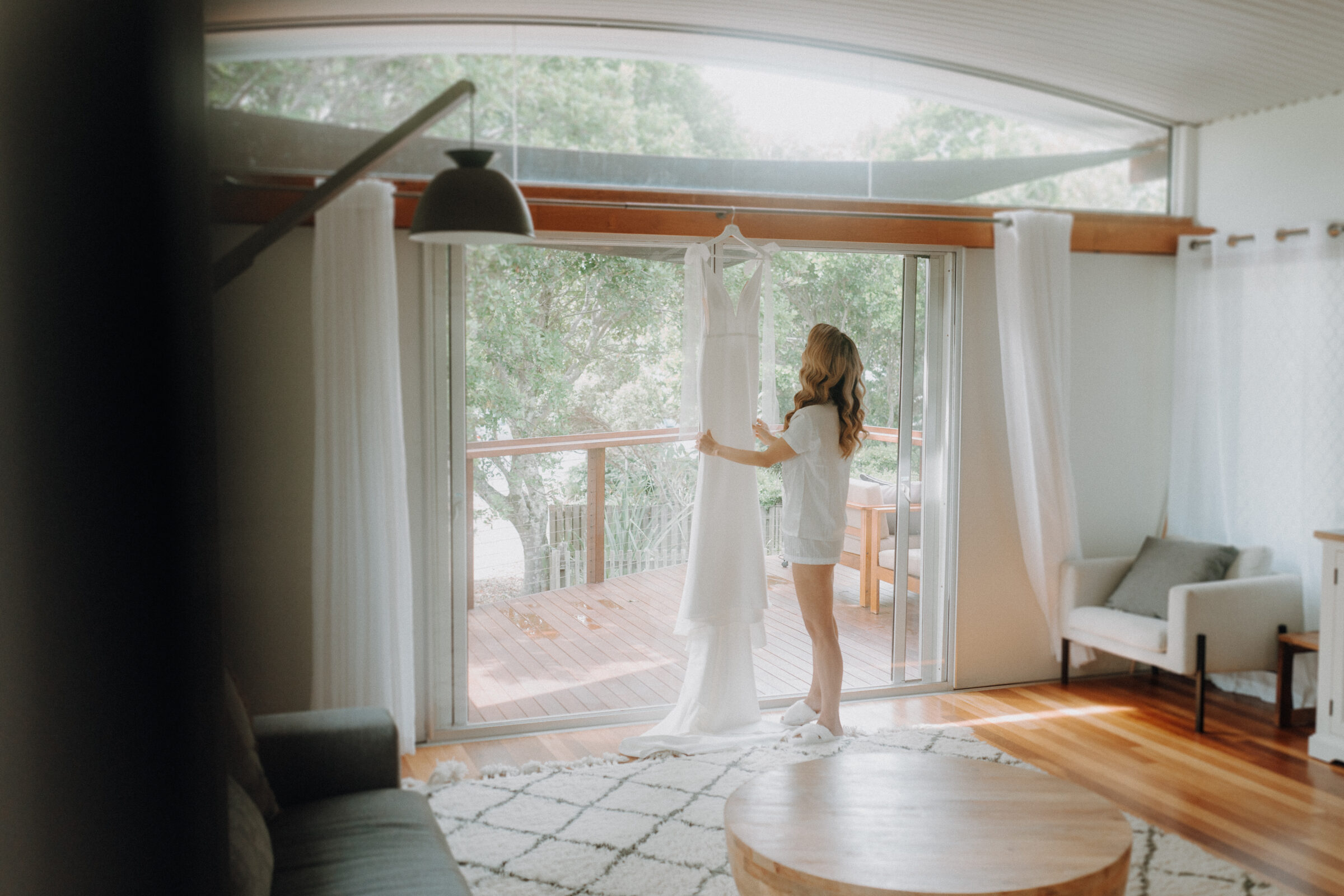 A woman stands in a bright room holding a white dress in front of a large window, overlooking a balcony with trees outside.