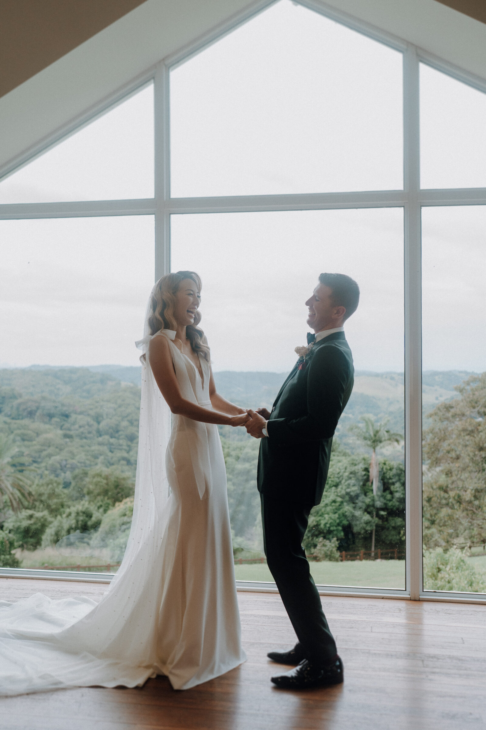 Bride in a flowing gown and groom in a black suit holding hands and smiling in front of a large window overlooking greenery.