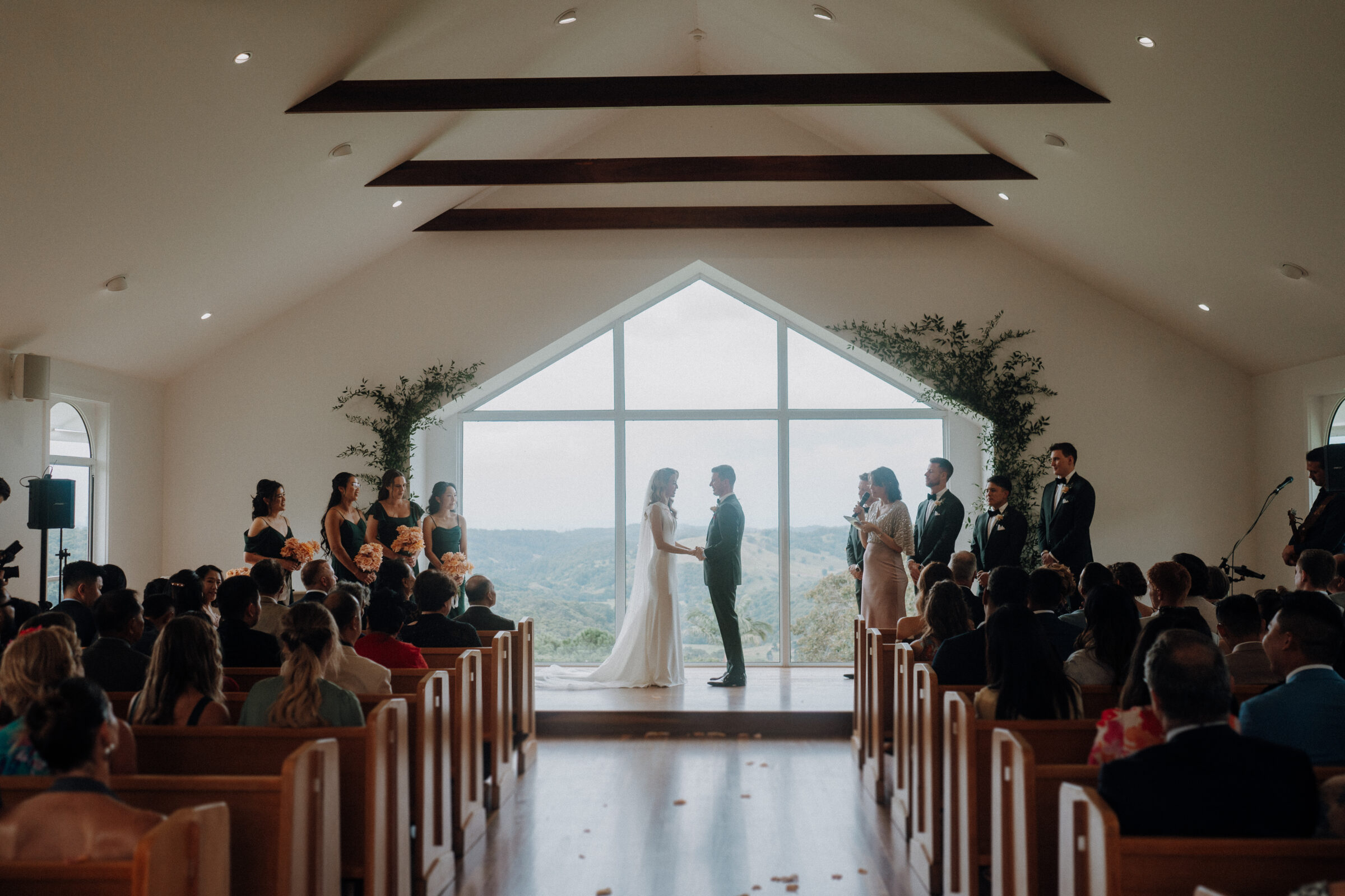 A couple stands at the altar in a church with large windows, surrounded by attendees and a wedding party.