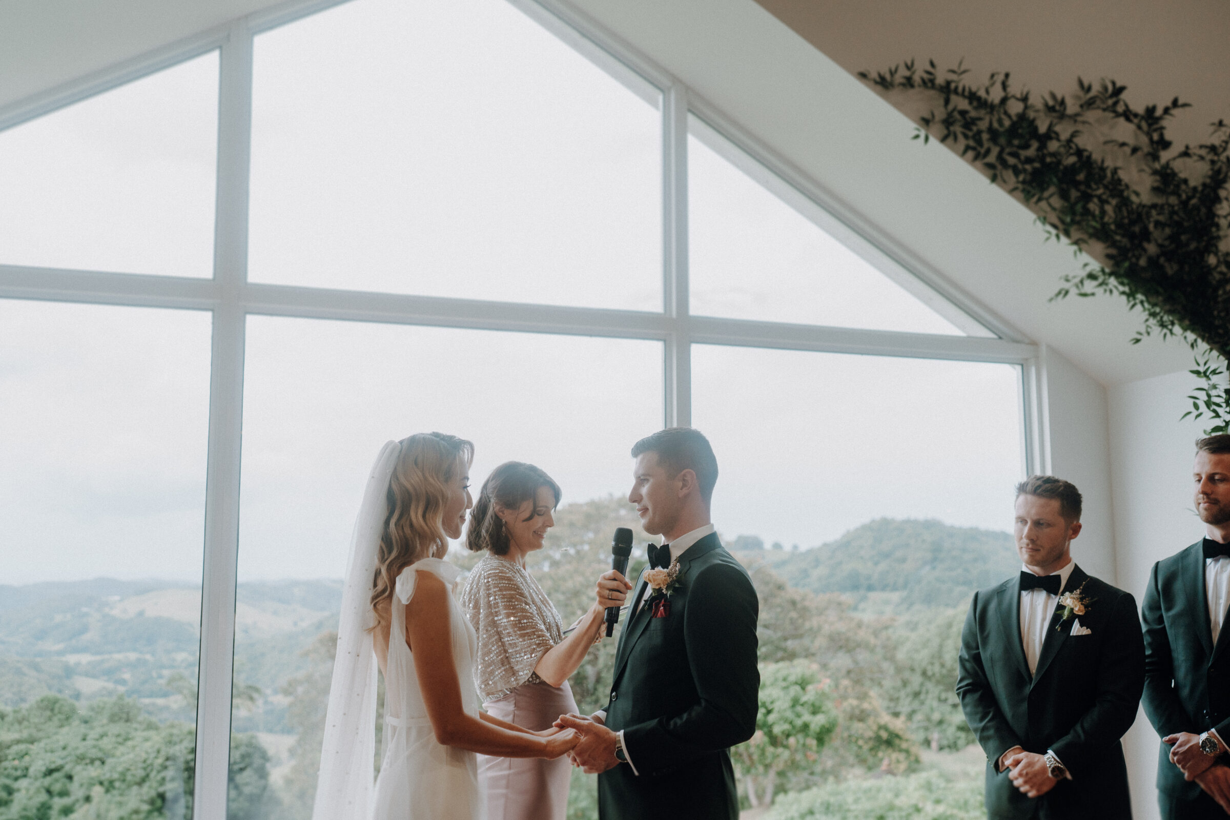 A bride and groom stand holding hands during a wedding ceremony in front of a large window with a scenic view. An officiant is speaking, while two groomsmen stand nearby.