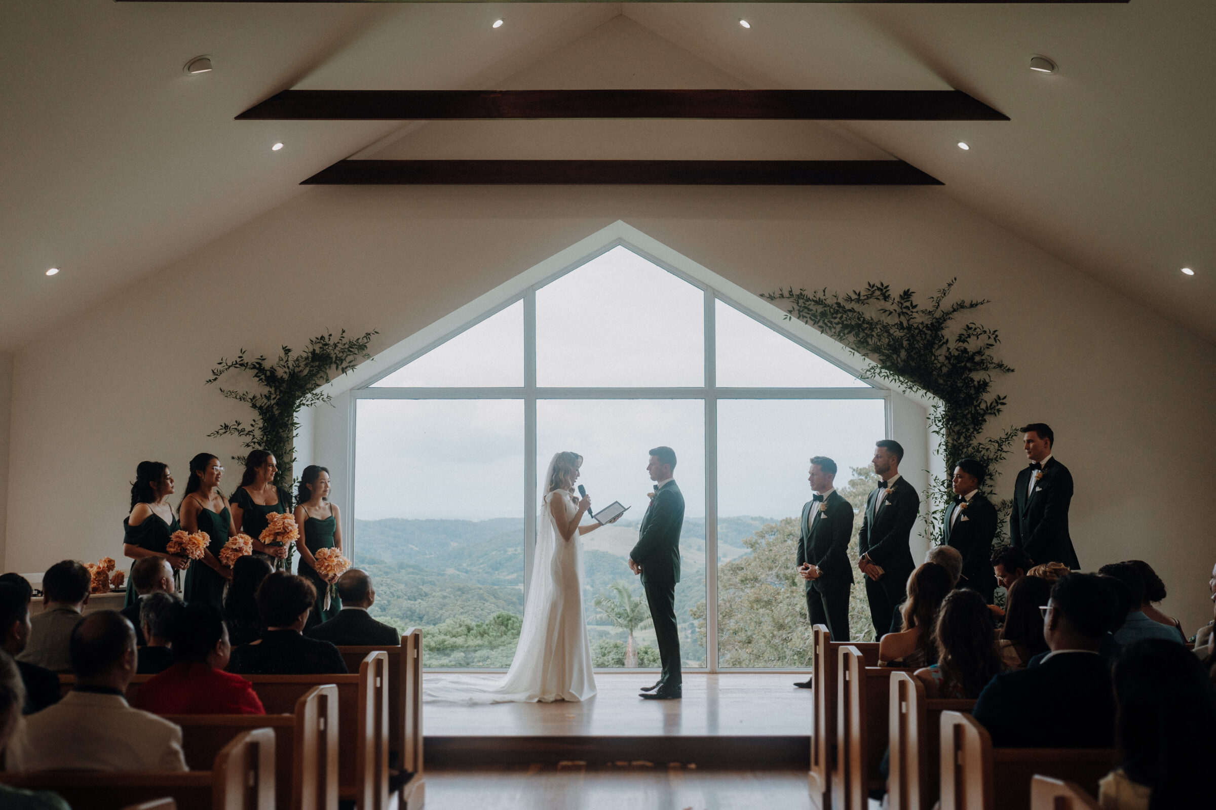 A couple stands at the altar exchanging vows in a chapel, surrounded by their bridal party and guests seated in pews. A large window showcases a scenic view of hills in the background.