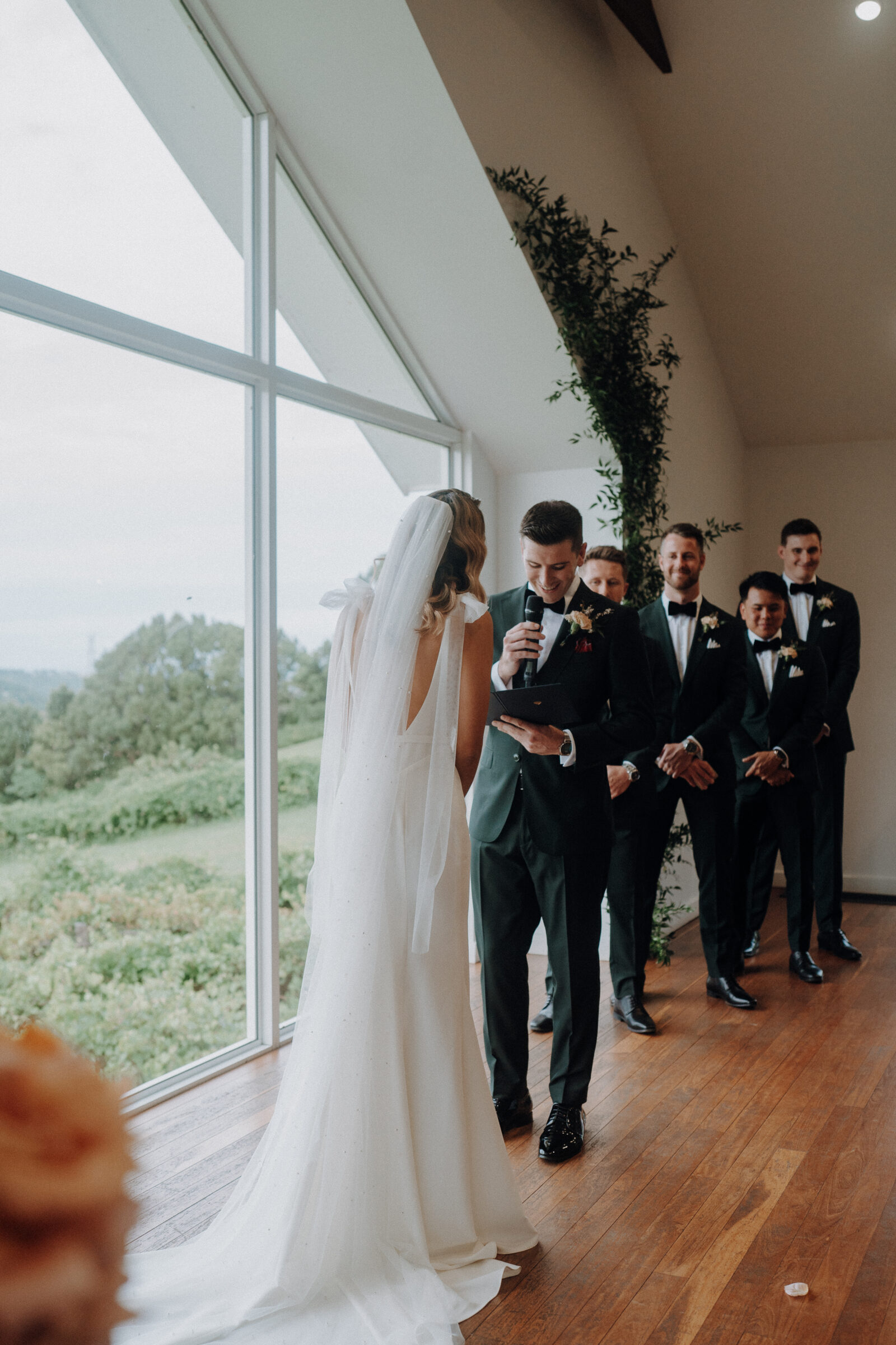 A bride and groom stand indoors near large windows. The groom is reading from a piece of paper. Four groomsmen stand behind them, smiling.