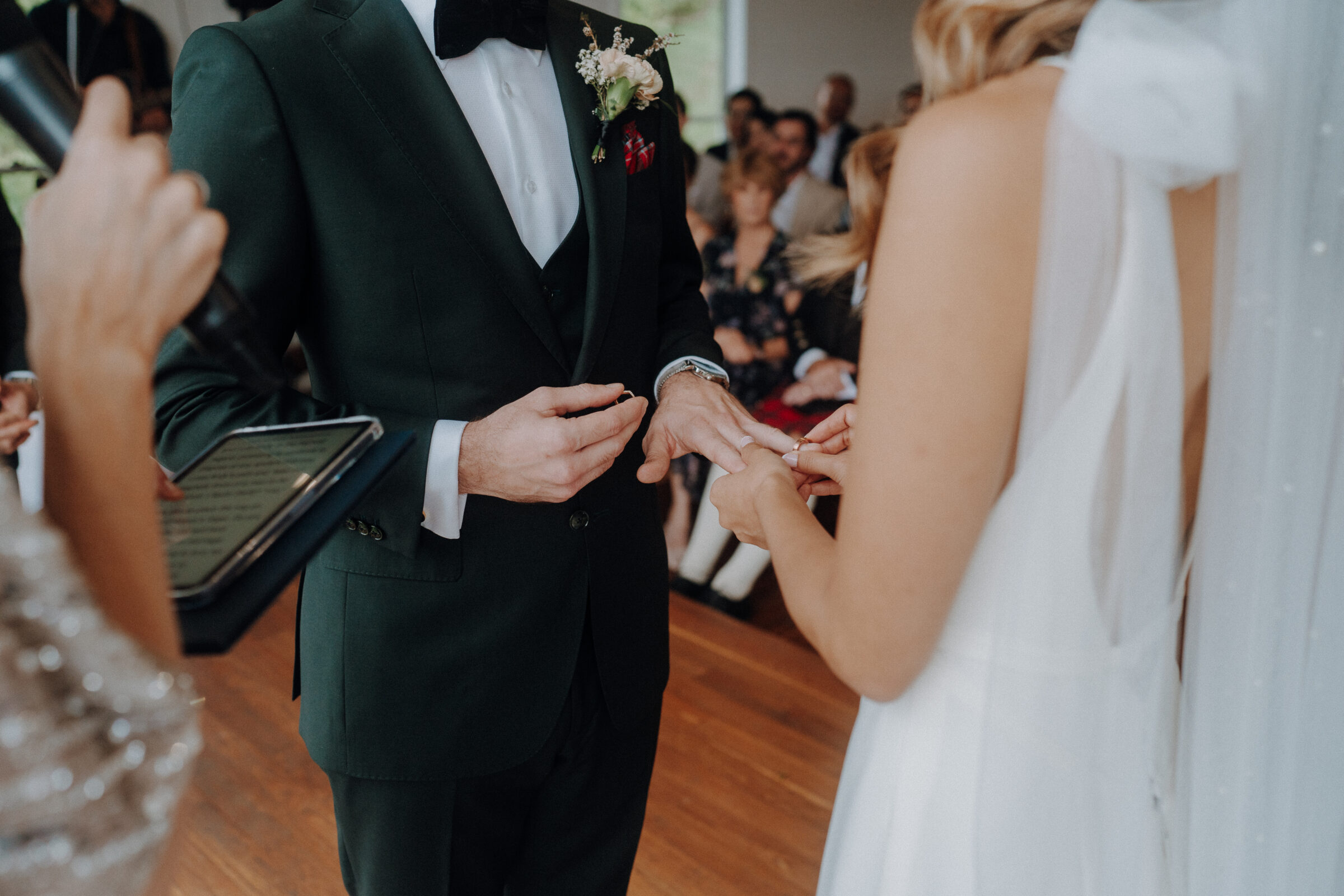 A bride and groom exchange rings during a wedding ceremony, with the officiant holding a microphone and a tablet. Guests are seated in the background.