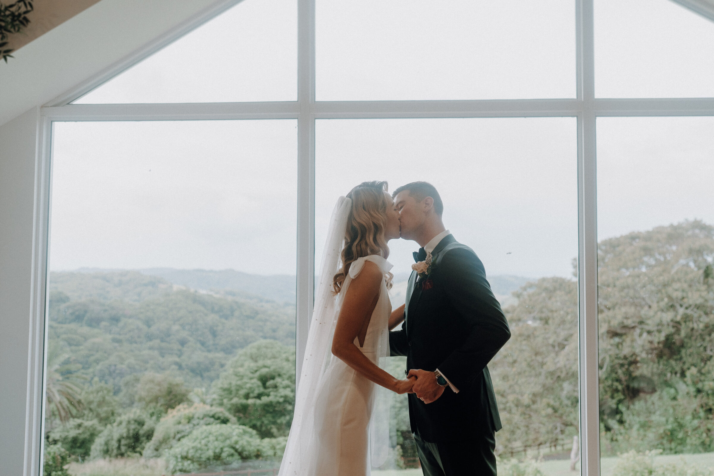 A bride and groom kiss in front of a large window with a view of a forested landscape in the background.