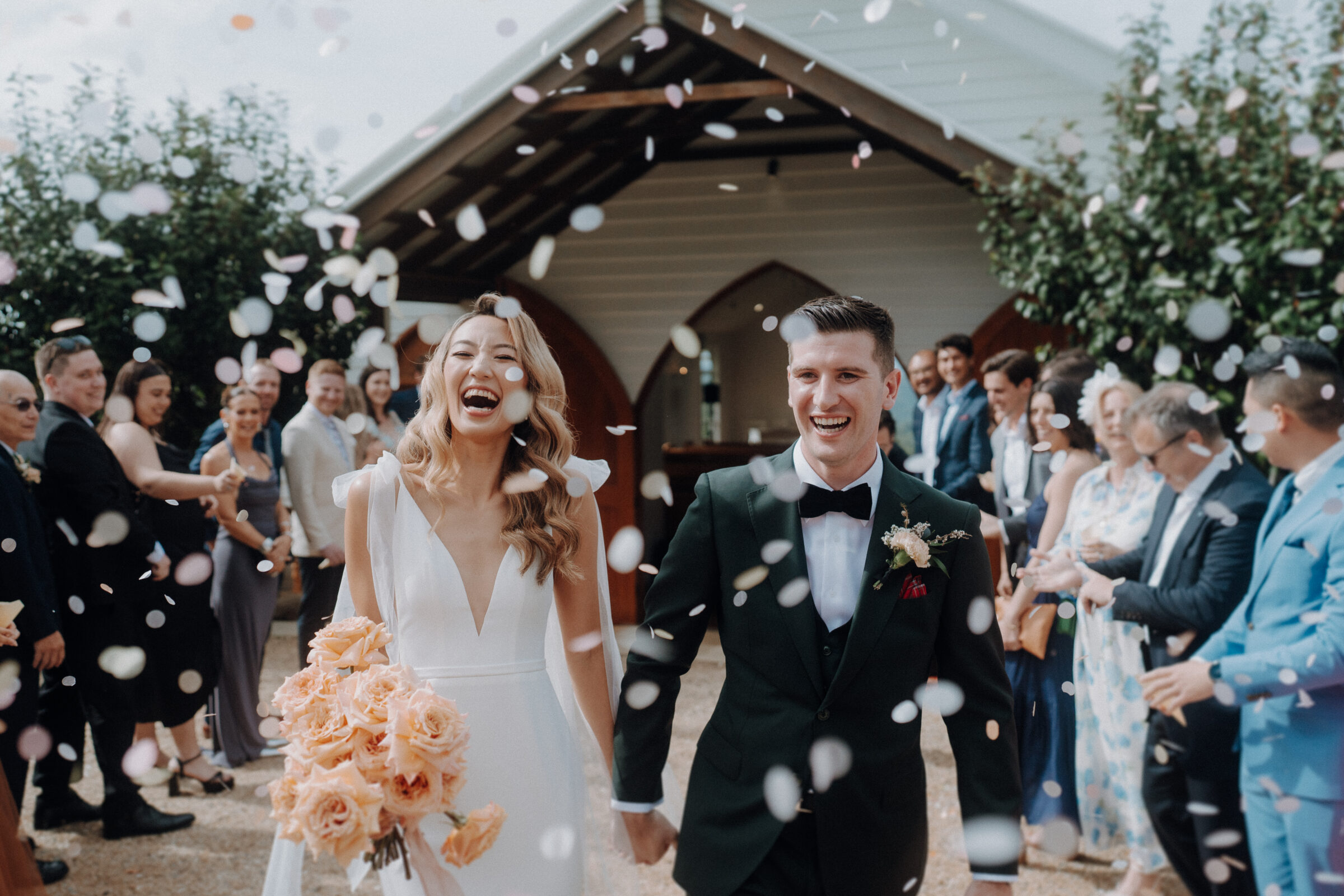 A couple in wedding attire walks through a crowd throwing flower petals outside a chapel. The bride holds a bouquet of roses, and guests are dressed in formal attire.