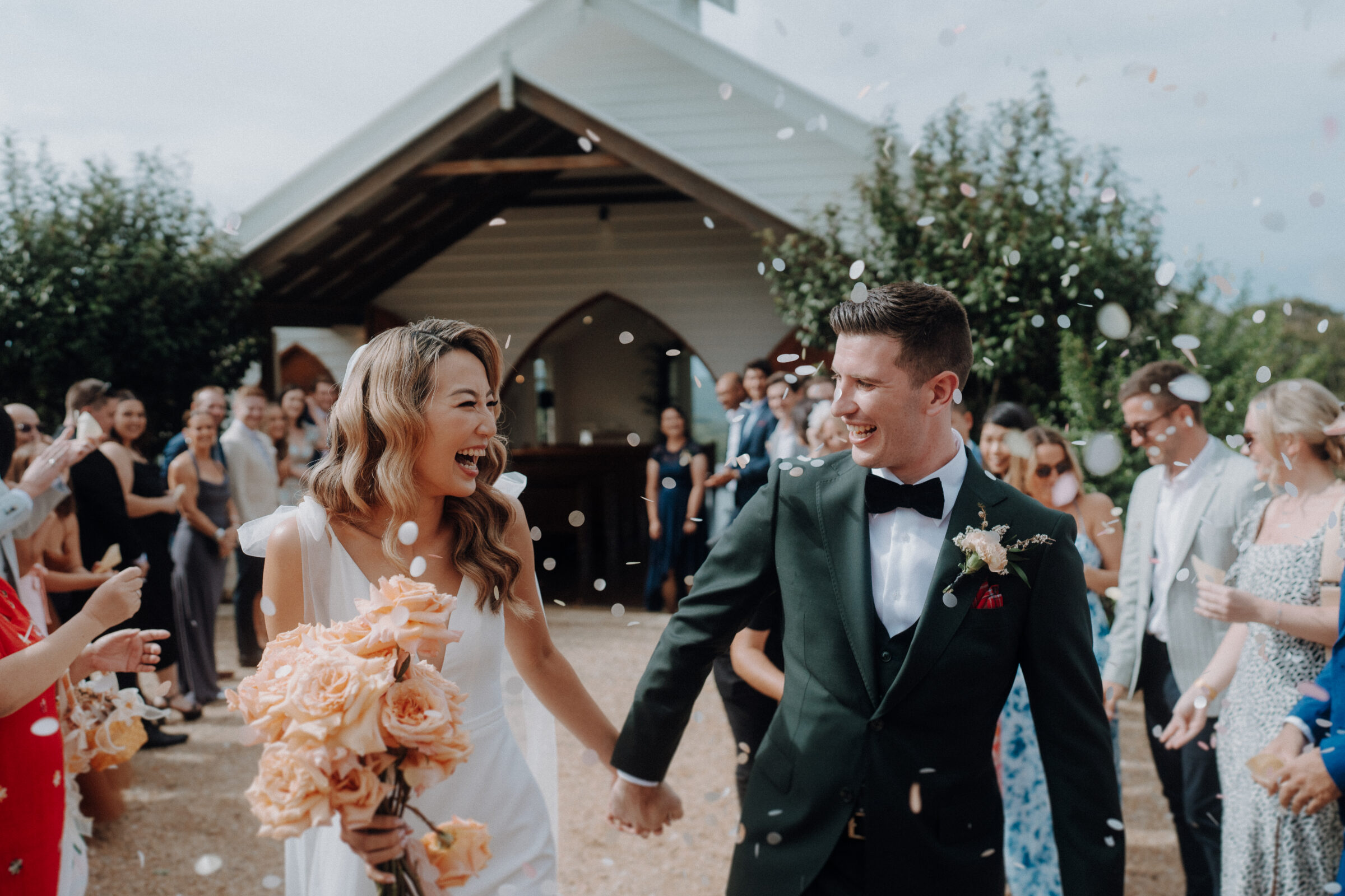 A newlywed couple exits a chapel surrounded by guests throwing confetti. The bride holds a bouquet of orange flowers.