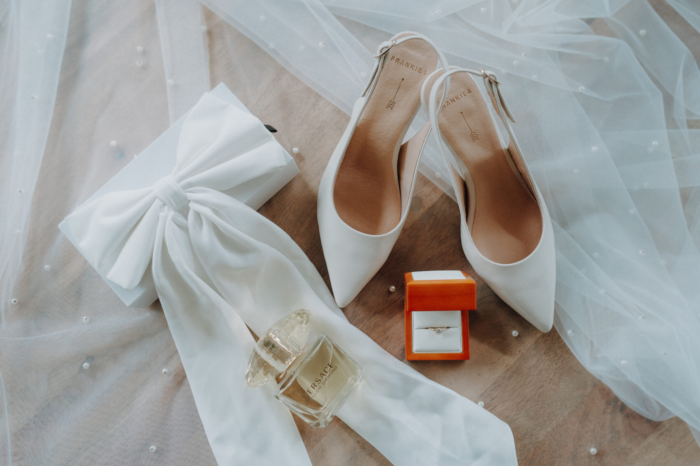 White bridal shoes and a matching clutch with a large bow are placed on sheer fabric, alongside a bottle of fragrance and an open jewelry box containing a ring.