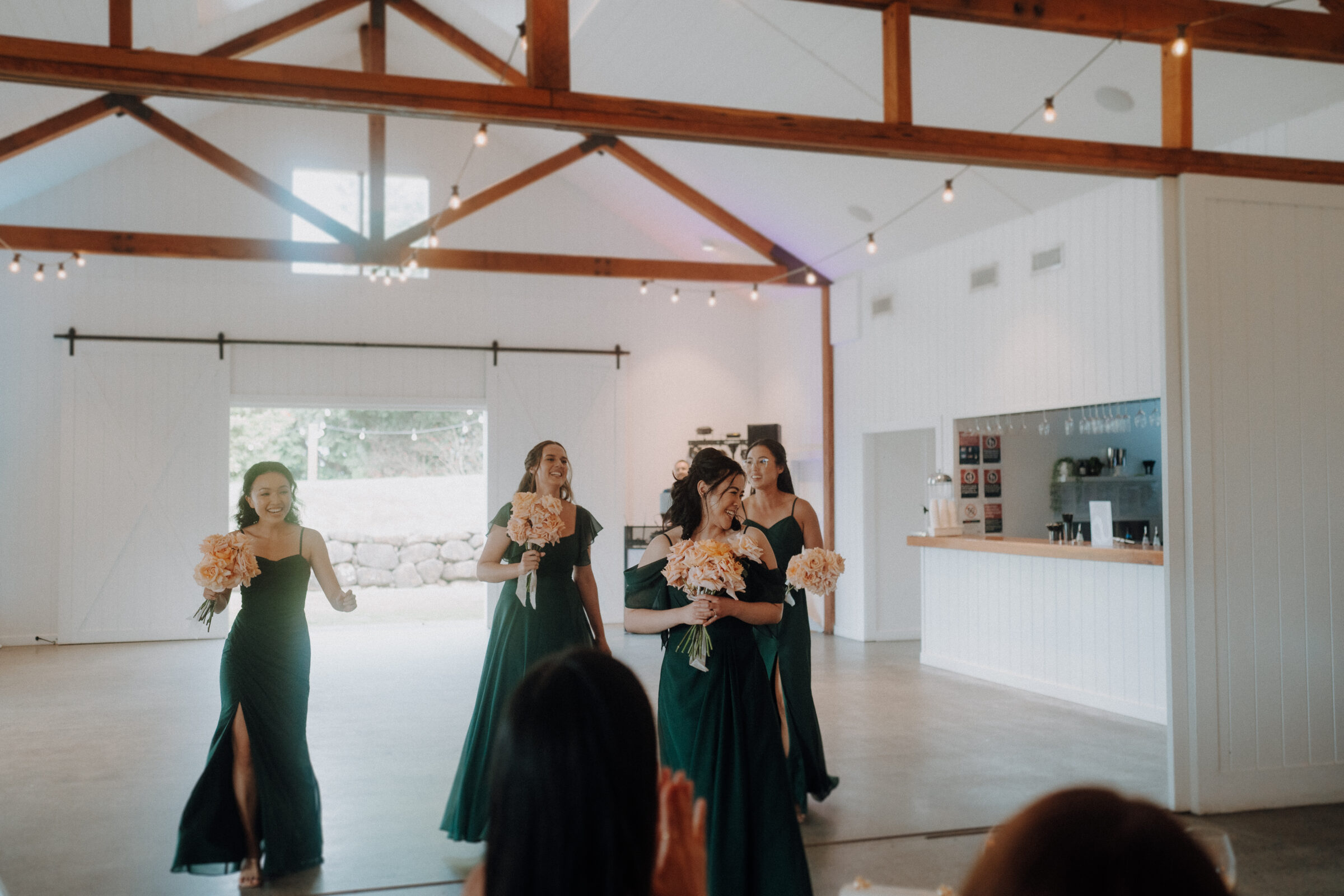 Five women in green dresses holding bouquets walk through a bright venue with exposed beams, while seated guests watch.