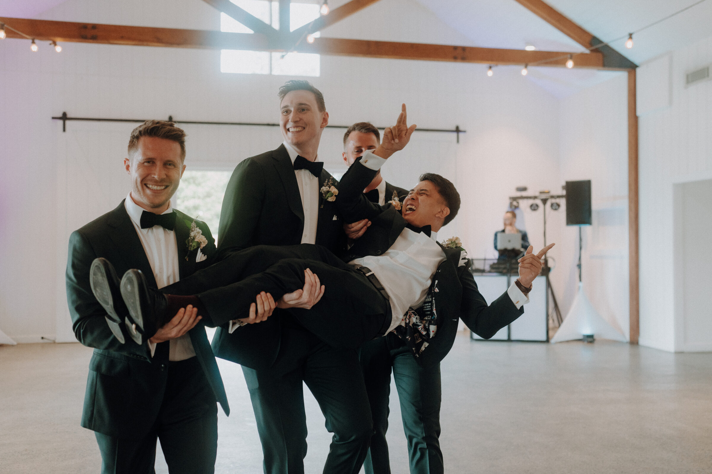 Three men in suits joyfully lift another man in a suit at an indoor event space. All are smiling.