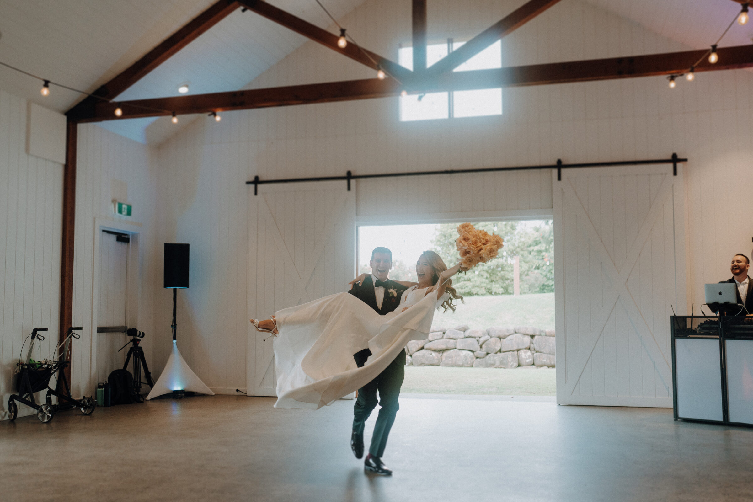 A groom spins a bride mid-air in a spacious, decorated barn with large windows and exposed beams.