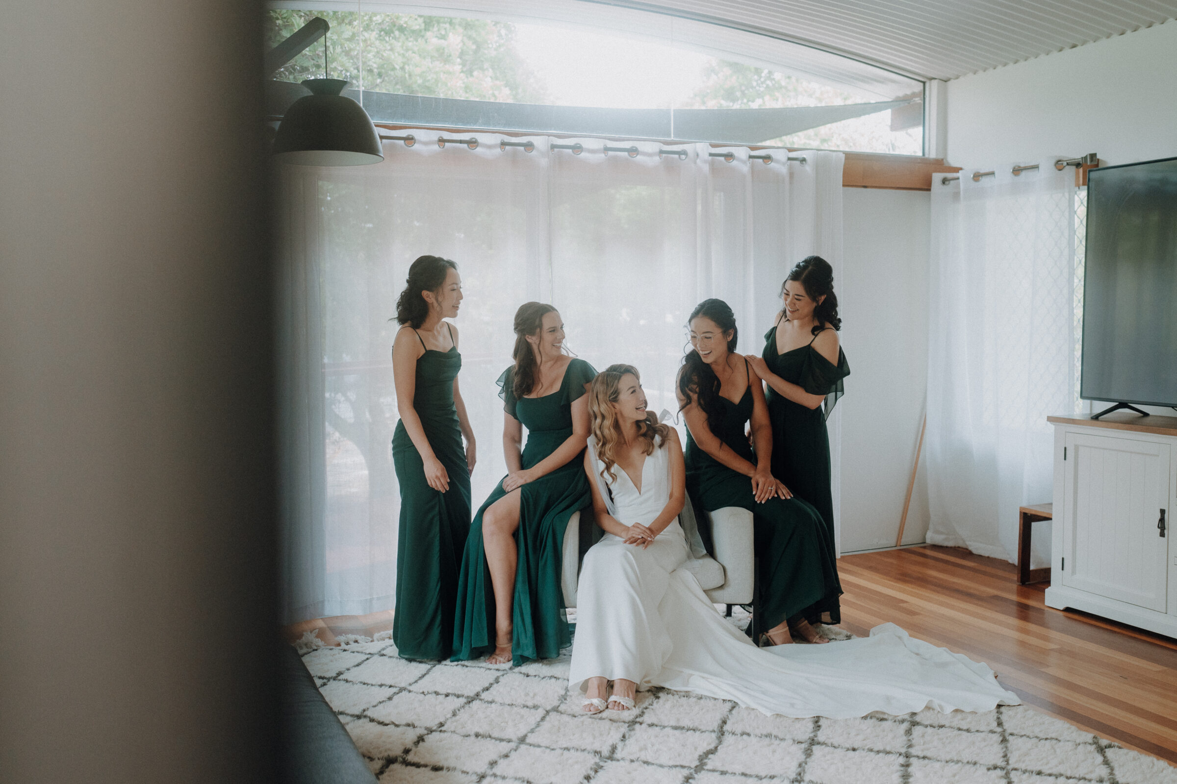 A bride sits in a white chair surrounded by bridesmaids in green dresses inside a room with large windows and sheer curtains.