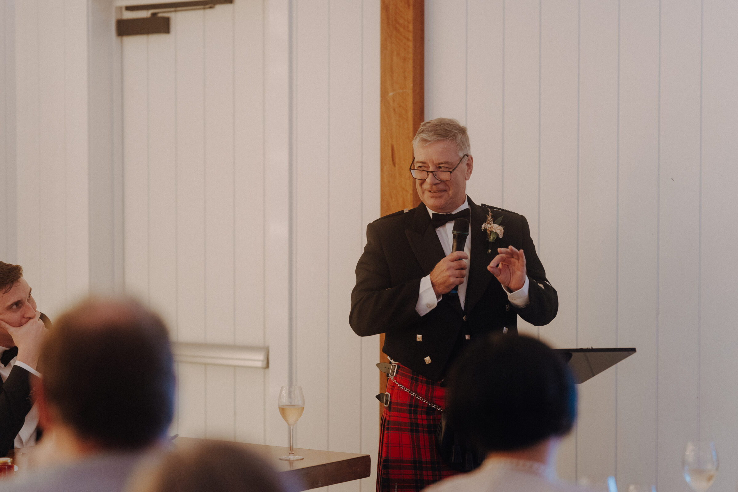 An older man in a suit and red tartan kilt speaks into a microphone at a lectern, in front of a white paneled wall, with glasses and attendees visible in the foreground.