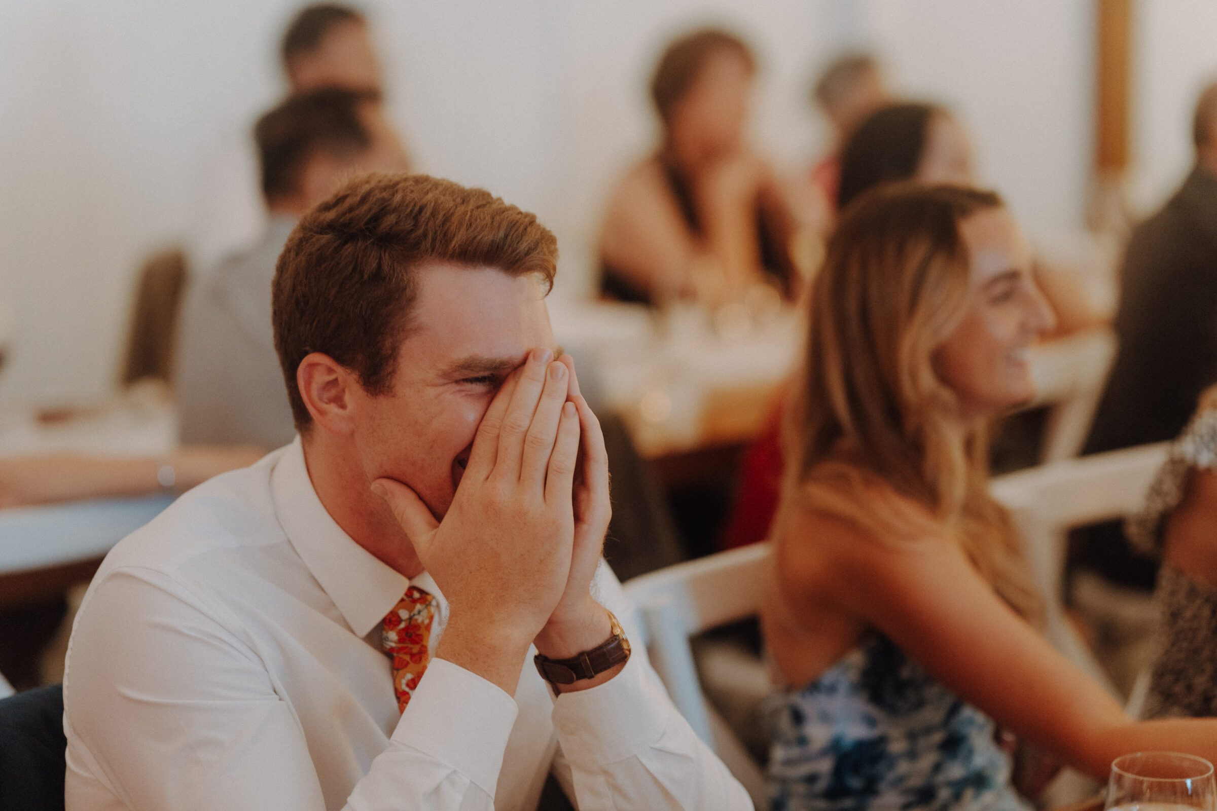 A man in a white shirt and patterned tie covers his face with his hands while laughing at an event, seated next to a woman in a patterned dress. Other attendees are blurred in the background.