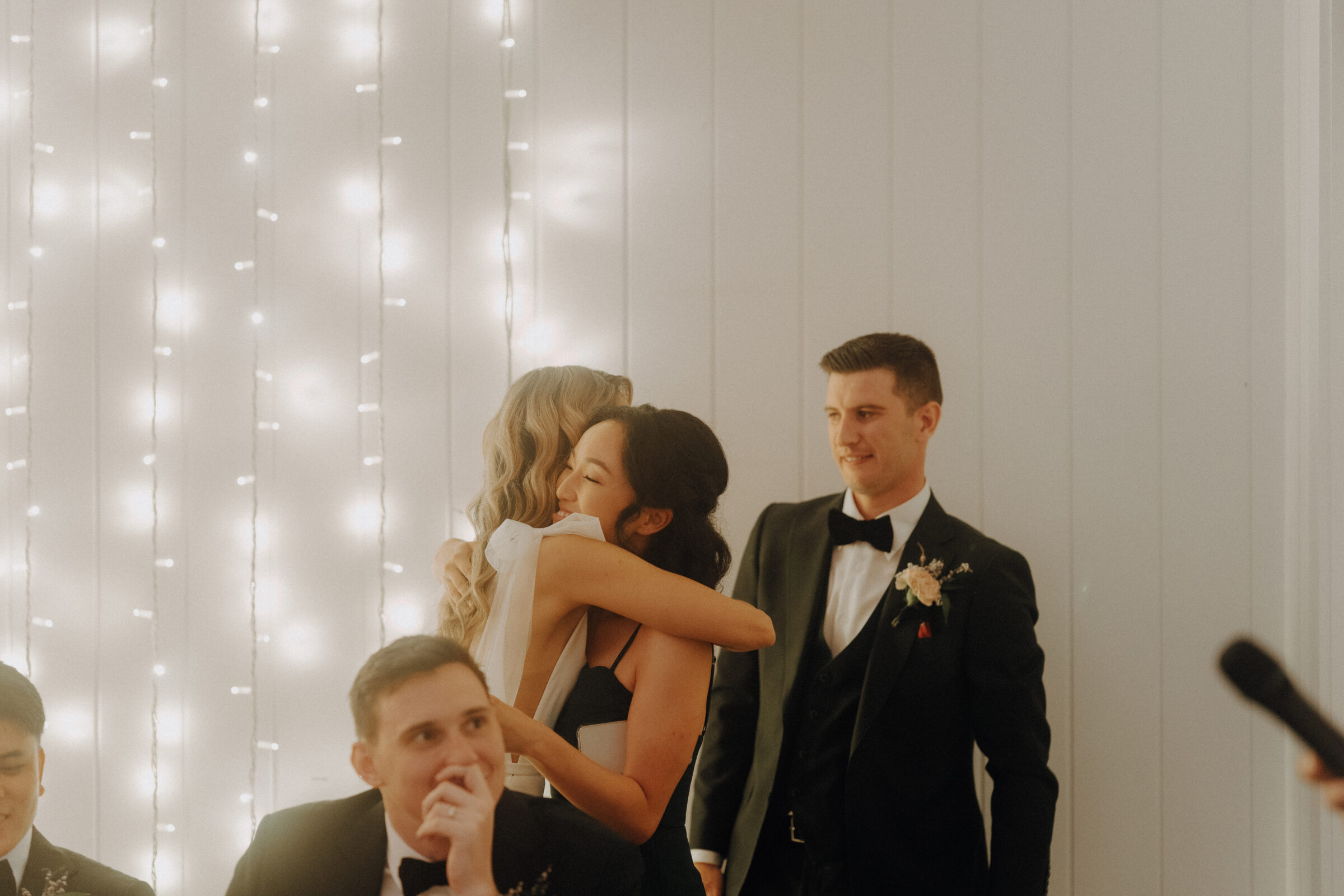 A woman hugs another woman in a black dress at an event, with a man in a suit standing nearby. String lights decorate the background.