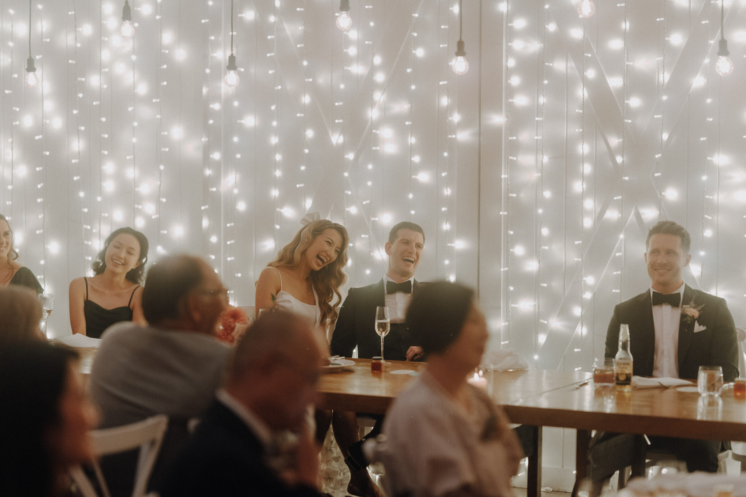 Guests at a wedding reception sitting at tables, smiling and laughing, with a backdrop of string lights.