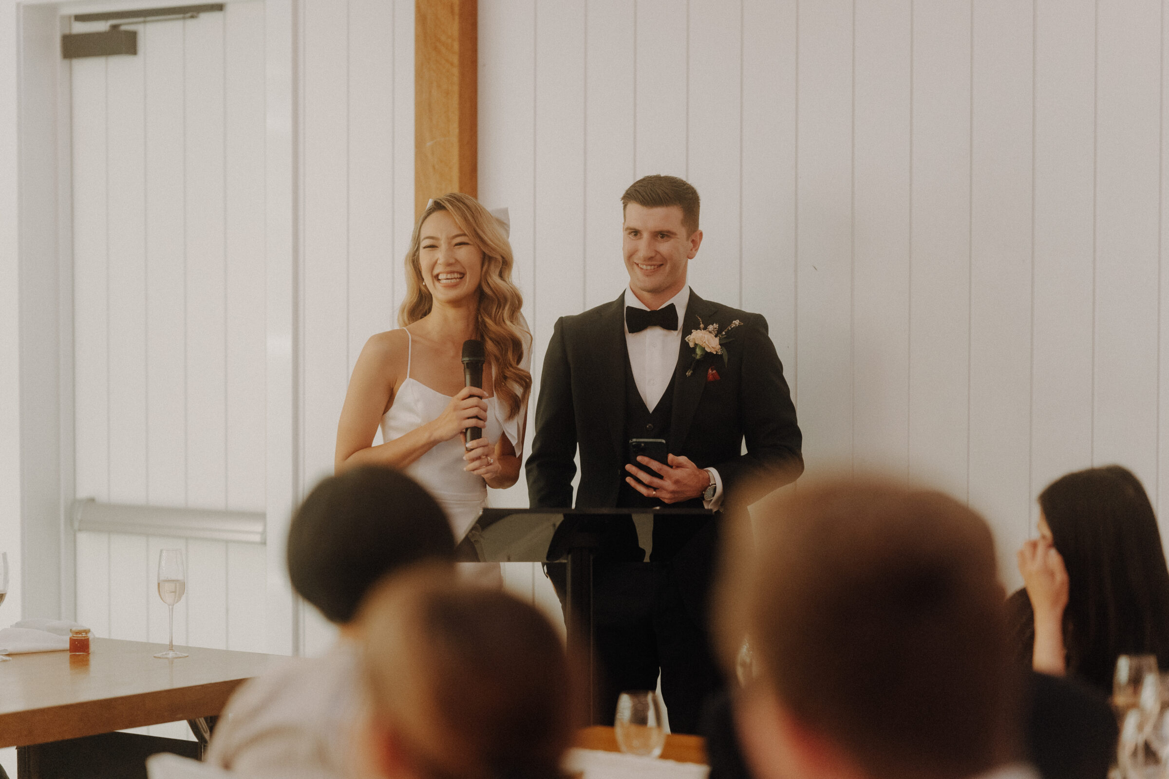 A bride and groom stand together at a microphone, smiling, in front of seated guests at a wedding reception.