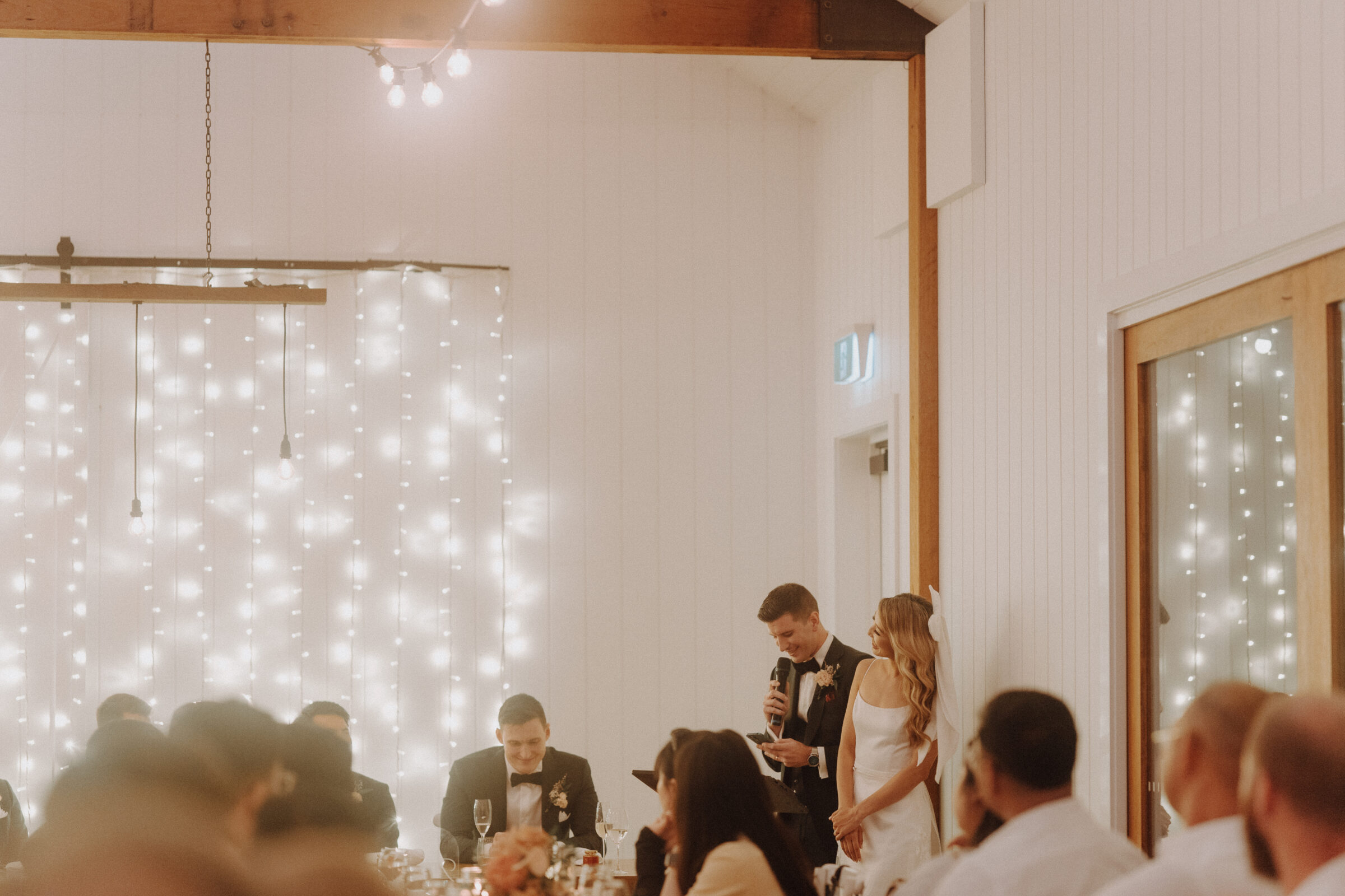 A bride and groom stand and speak at a wedding reception in a warmly lit room with guests seated at tables and fairy lights in the background.