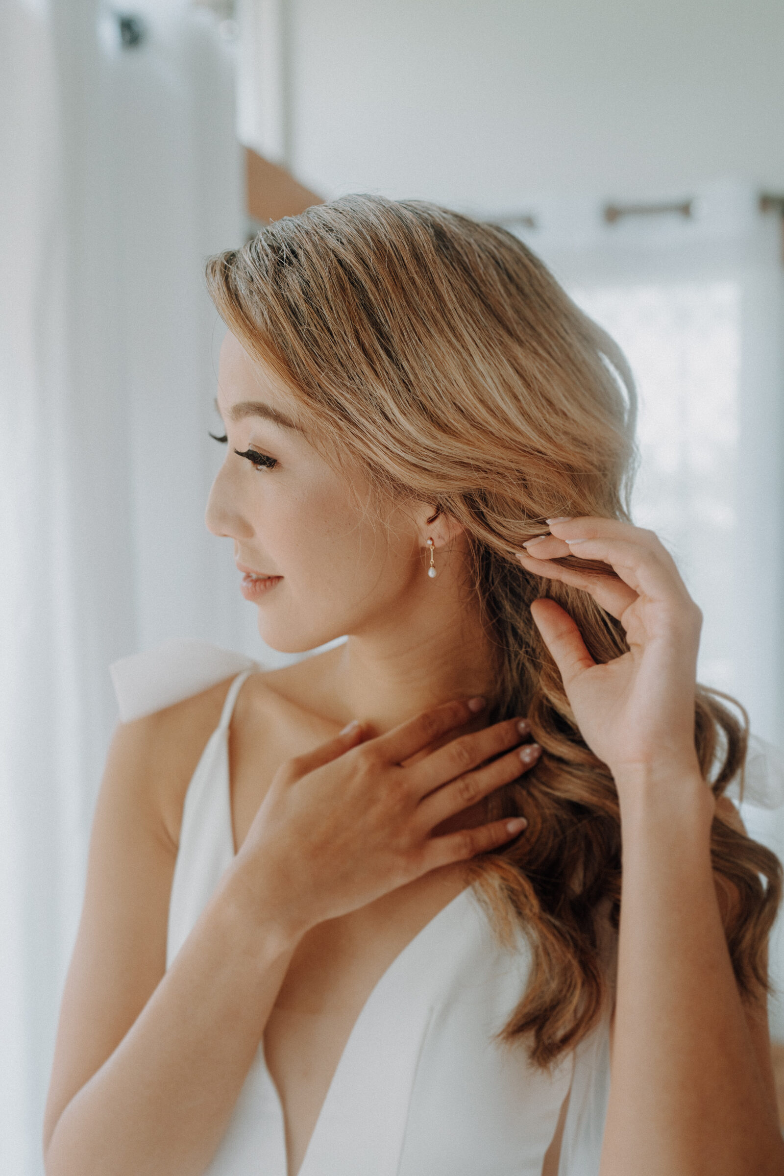 A woman in a white dress stands by a window, touching her long, wavy hair with one hand and resting the other on her chest. She gazes off to the side.