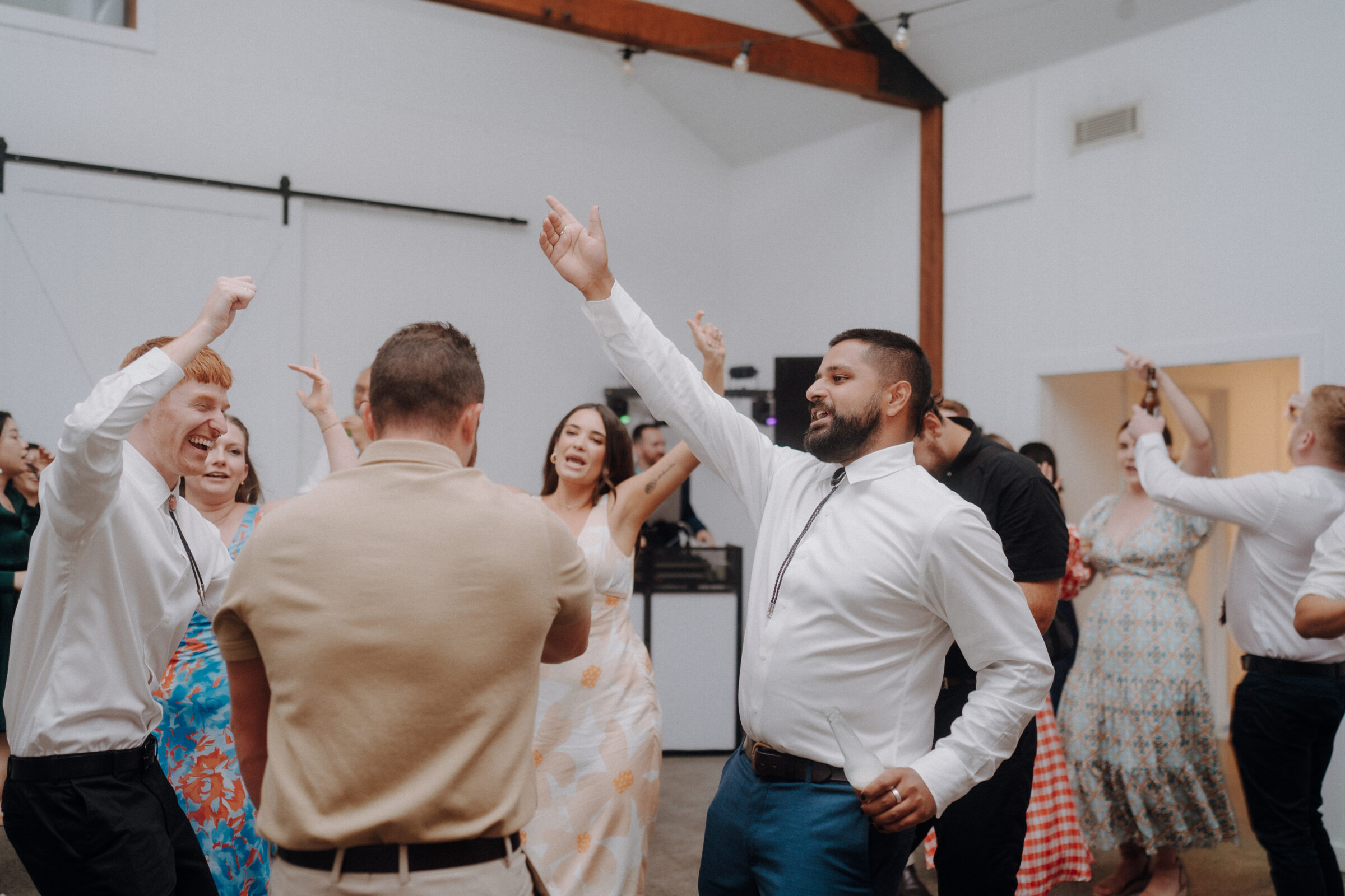 People dancing energetically in a white room with wooden beams, wearing casual and semi-formal attire.