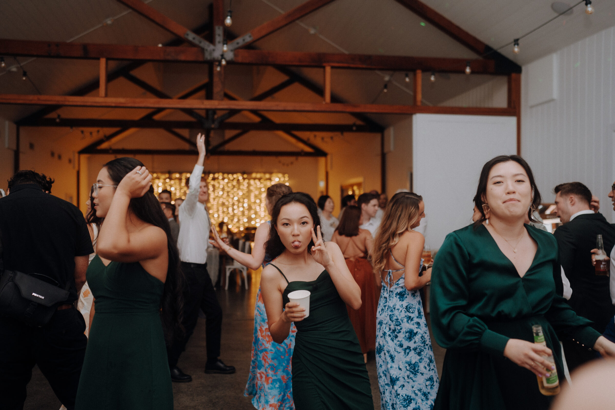 People dancing and mingling at an indoor event with a decorated backdrop. One person makes a peace sign gesture while holding a cup.