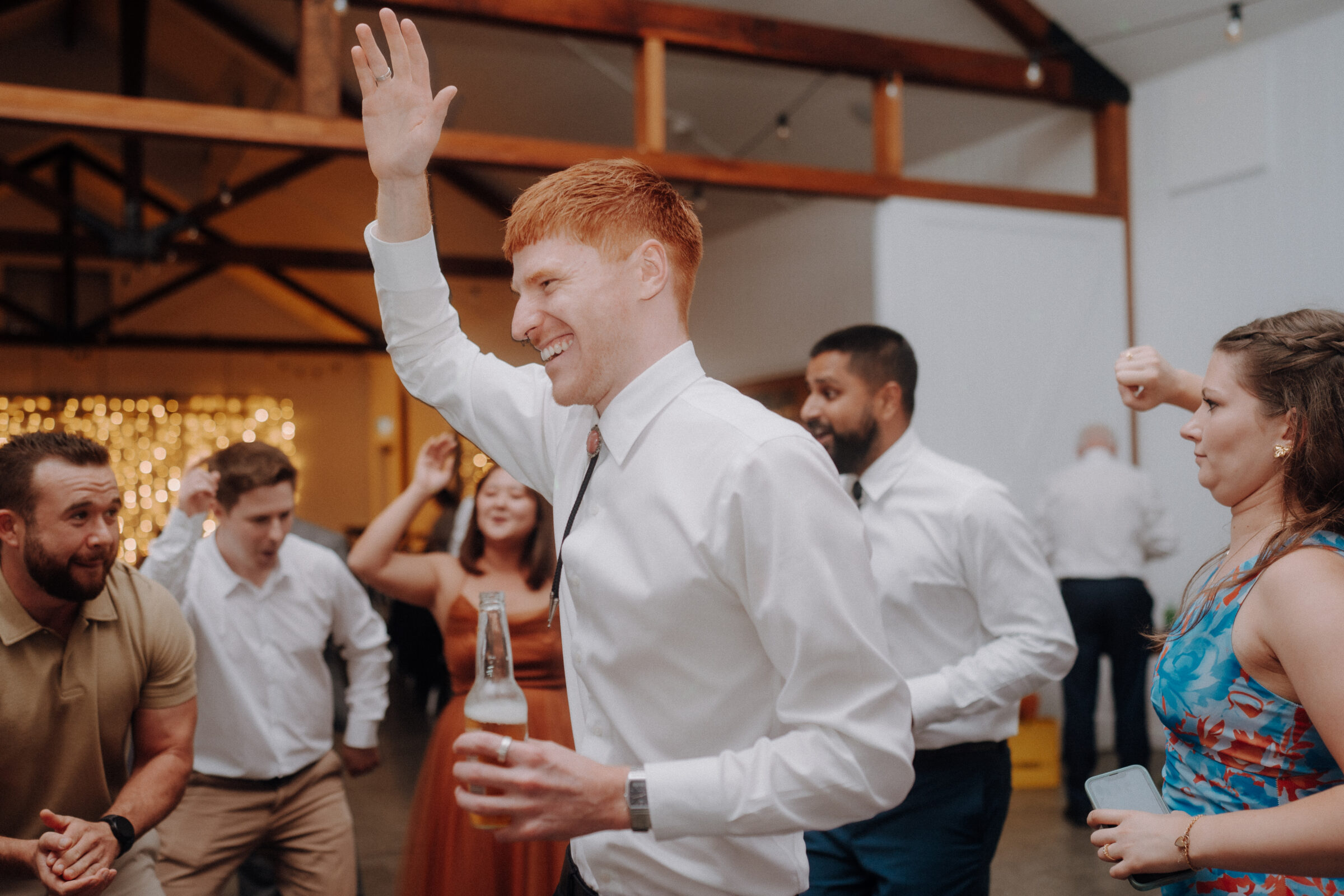 A group of people dancing at an indoor event. A man in a white shirt holds a bottle and raises his hand.