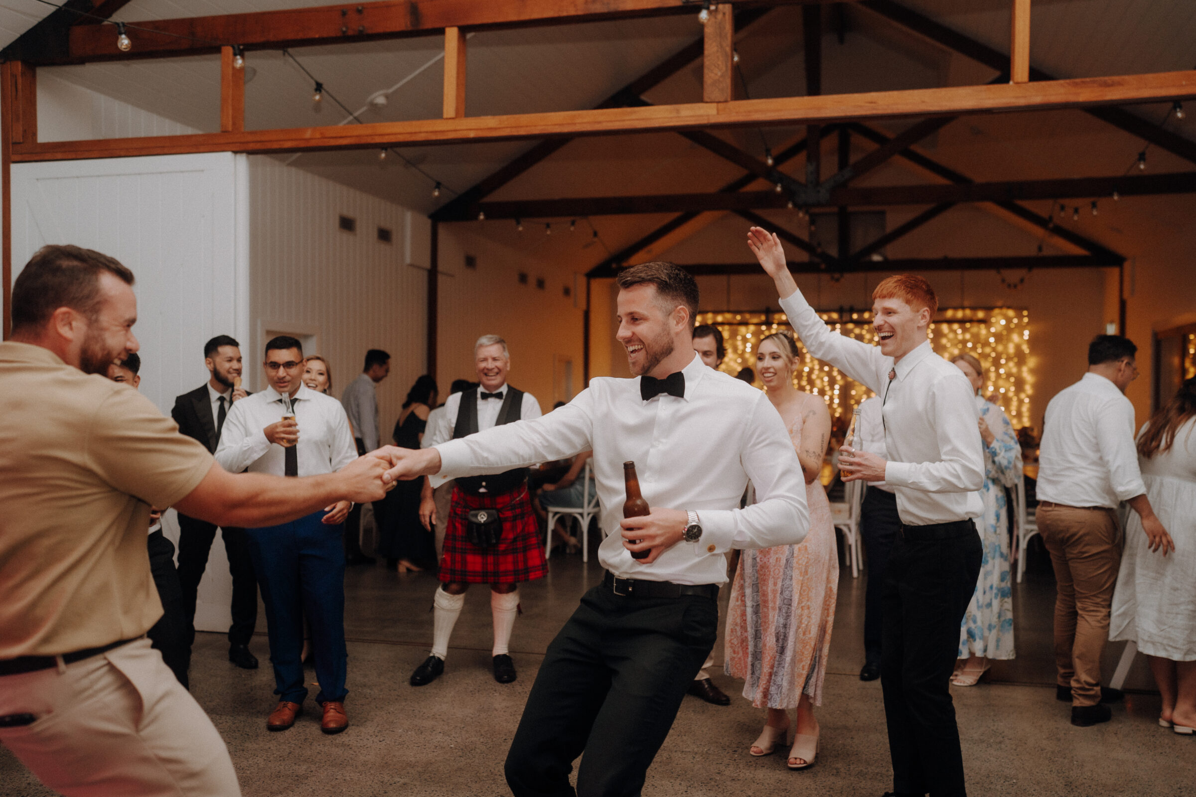 People are dancing and socializing at an indoor event, with festive lights in the background. One man is wearing a kilt.