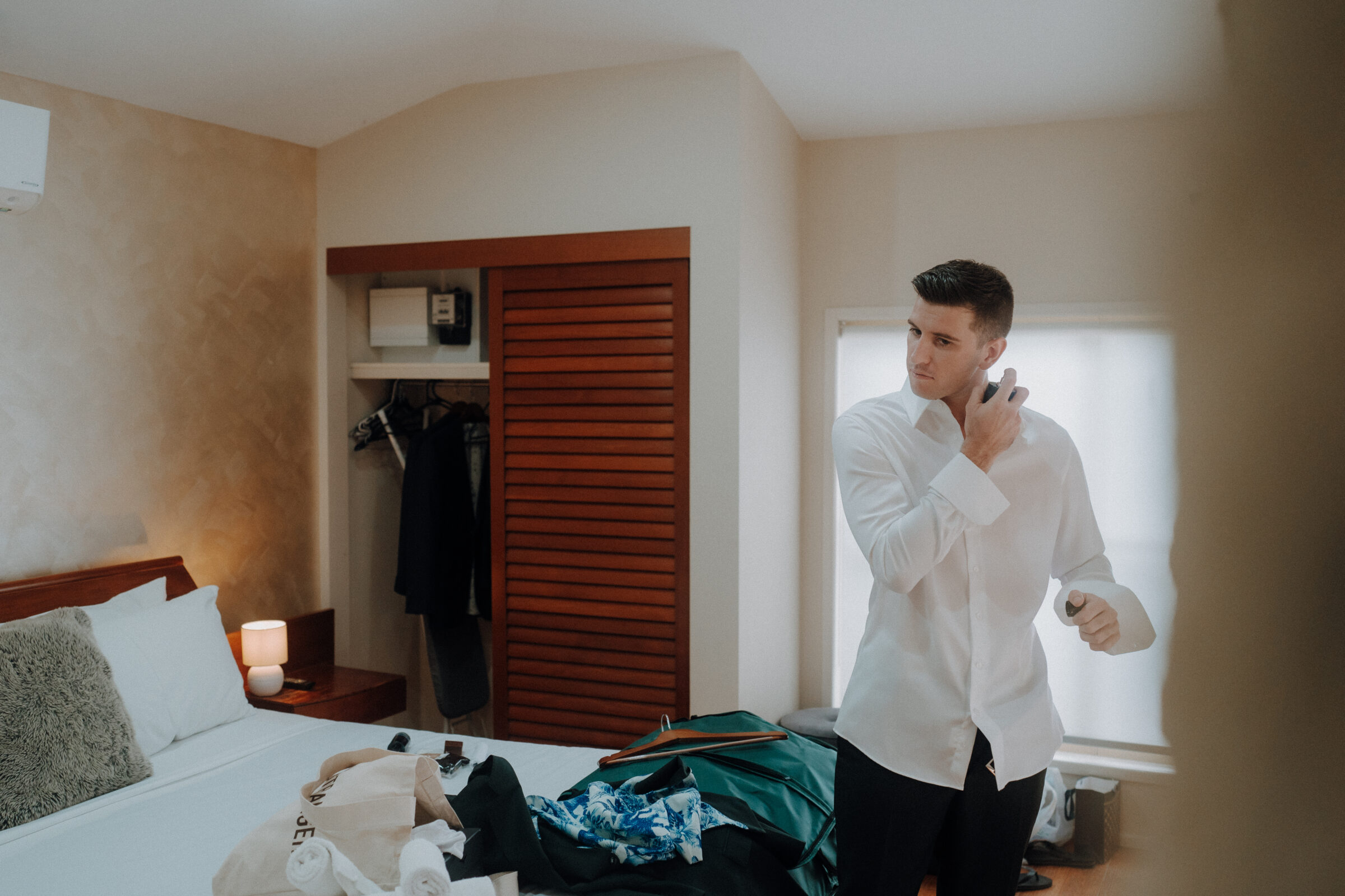 A man in a white shirt applies cologne in a bedroom with an unmade bed, clothes, and a teal suitcase scattered around.