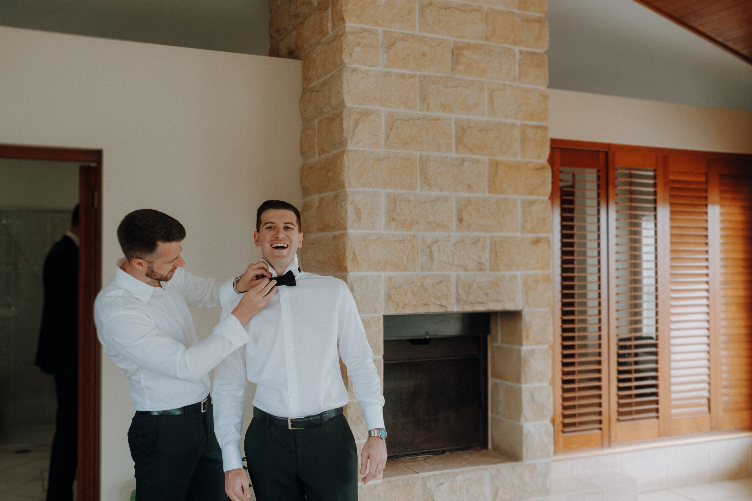 Two people in formal attire; one is helping the other with a bow tie in a room with a stone fireplace and wooden shutters.