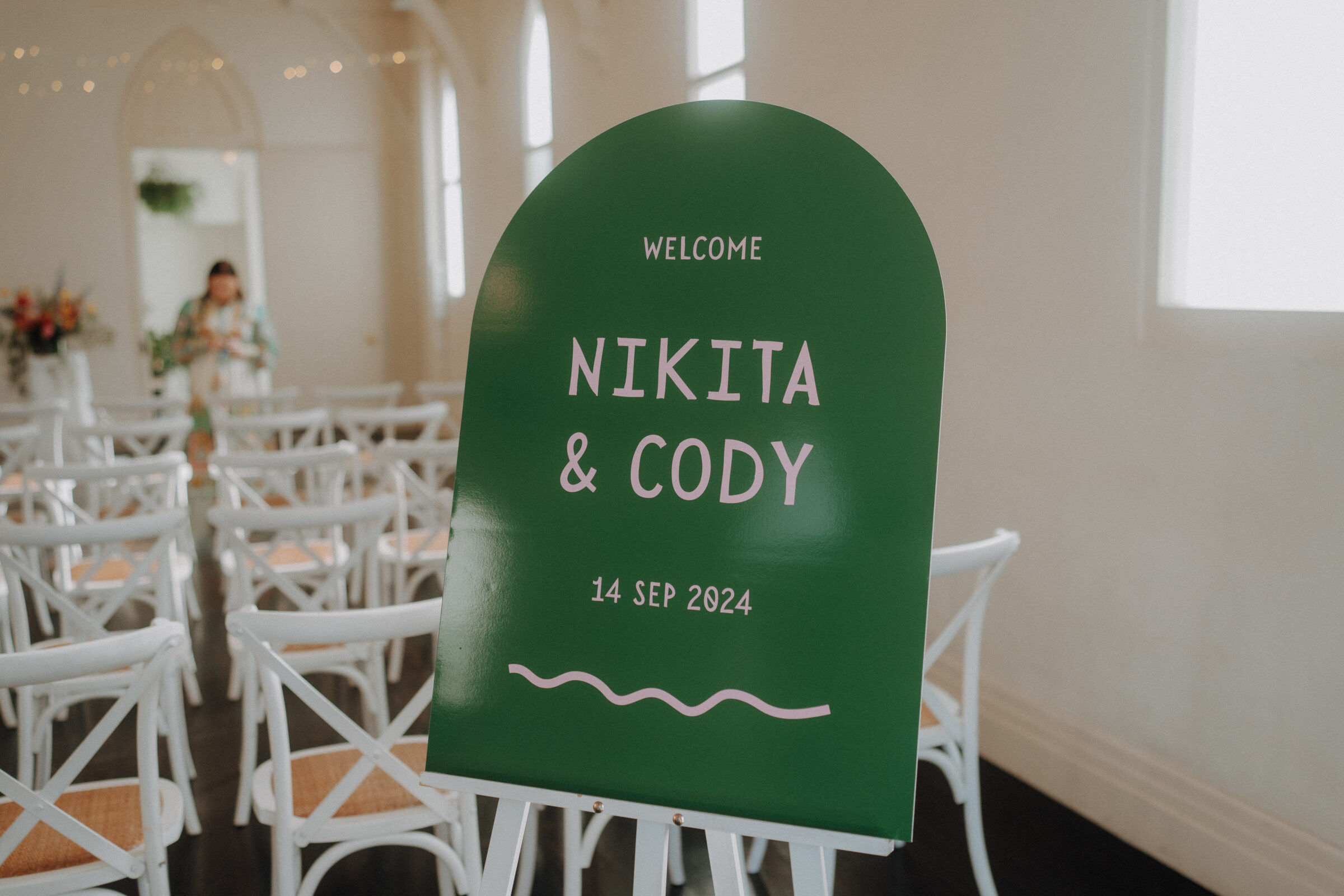 Green sign with "Welcome Nikita & Cody, 14 Sep 2024" in a room with white chairs and a person in the background.