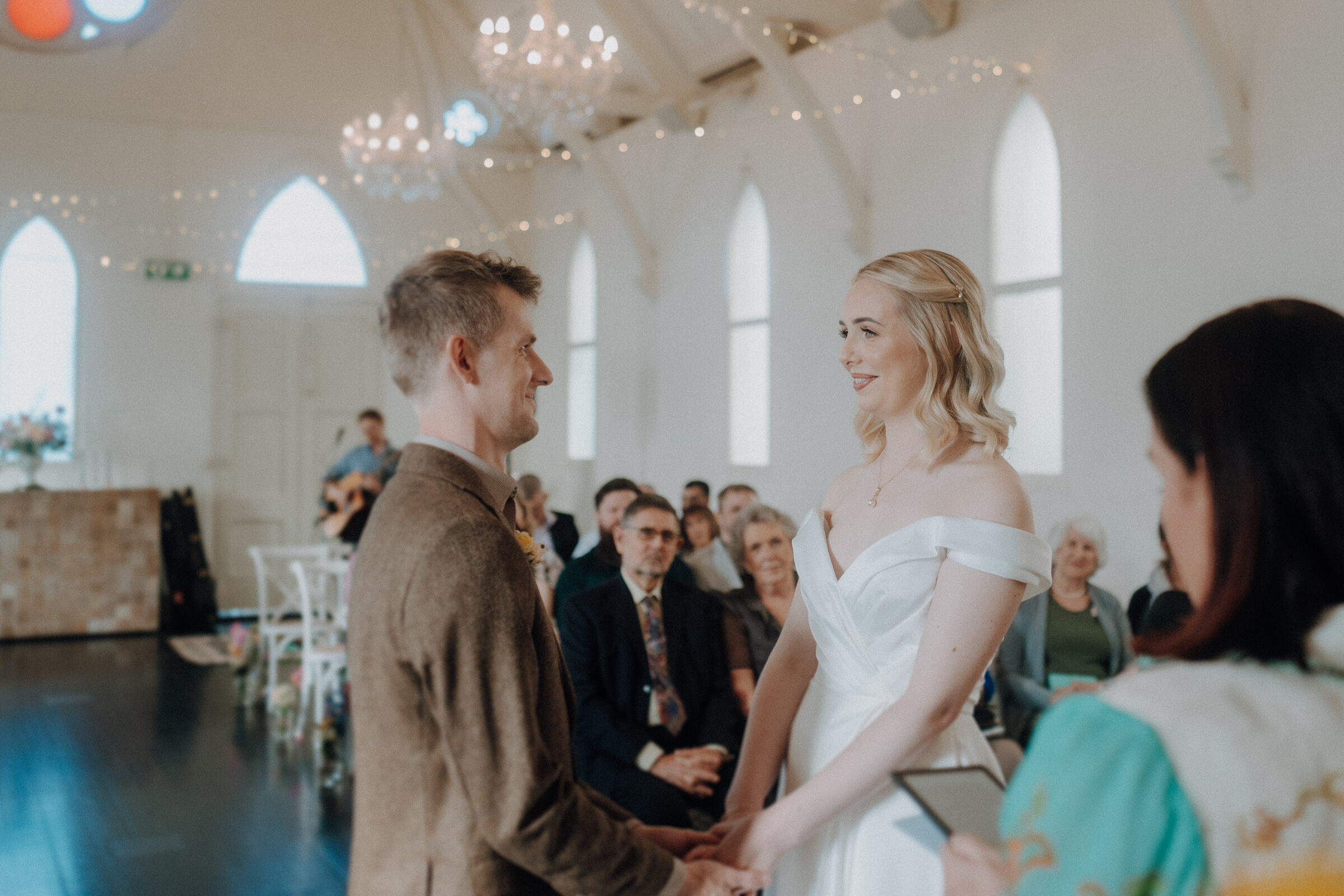 A couple in formal attire stands facing each other, holding hands, at a wedding ceremony with guests seated in the background.