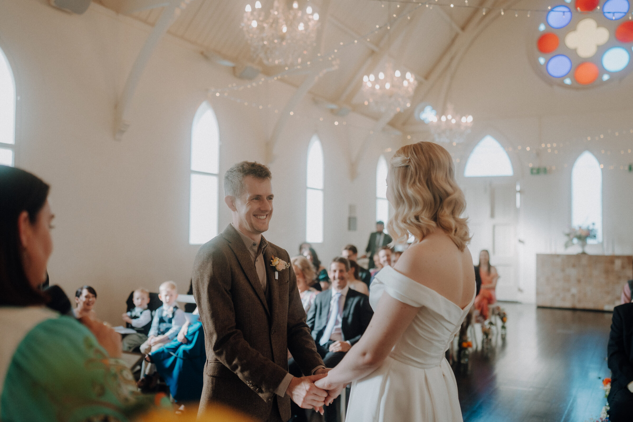 A couple stands at the altar in a bright church, exchanging vows. Guests are seated, watching the ceremony. Elegant chandeliers and stained glass are visible.