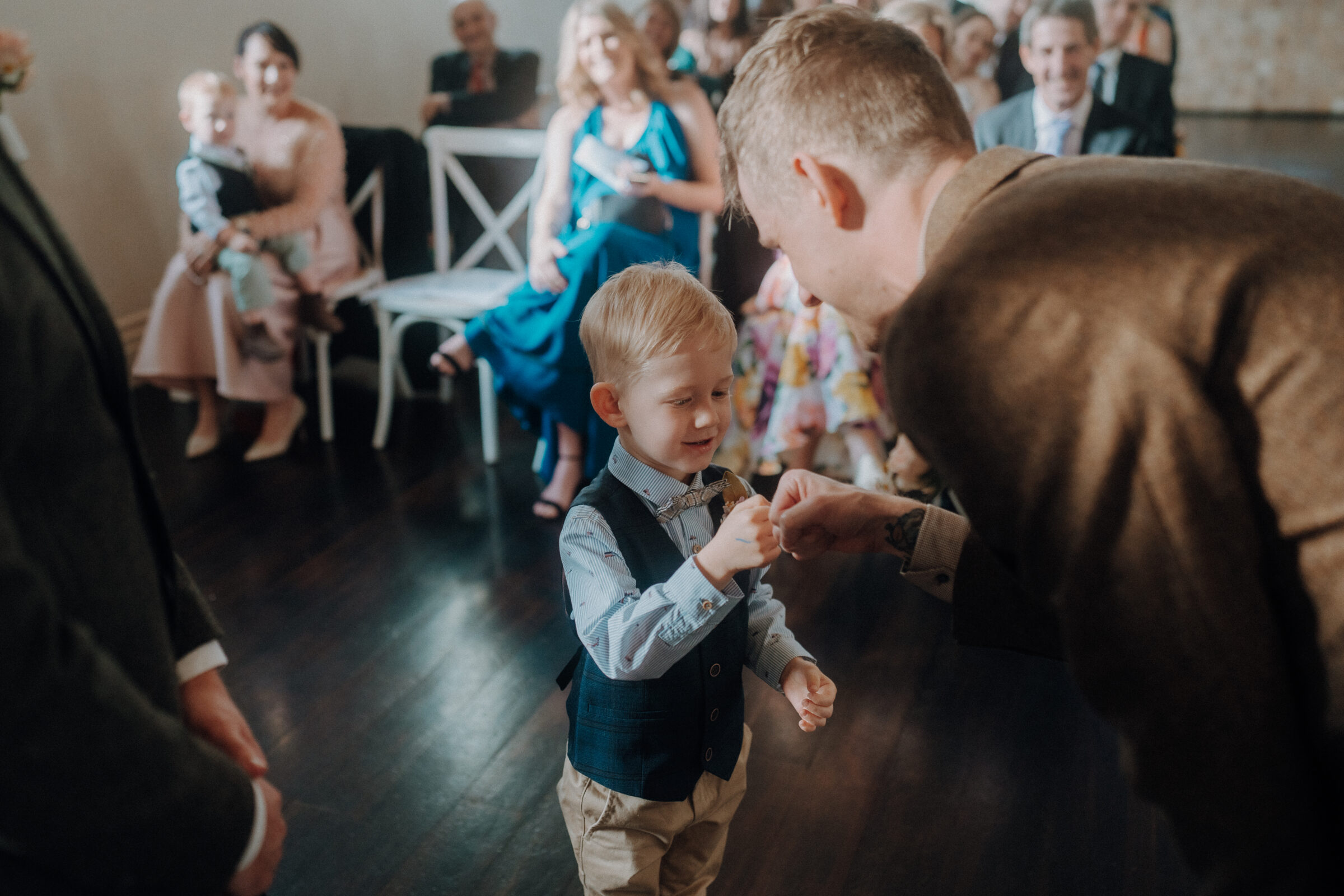 A young boy in formal attire high-fives an adult at an indoor event with seated guests in the background.