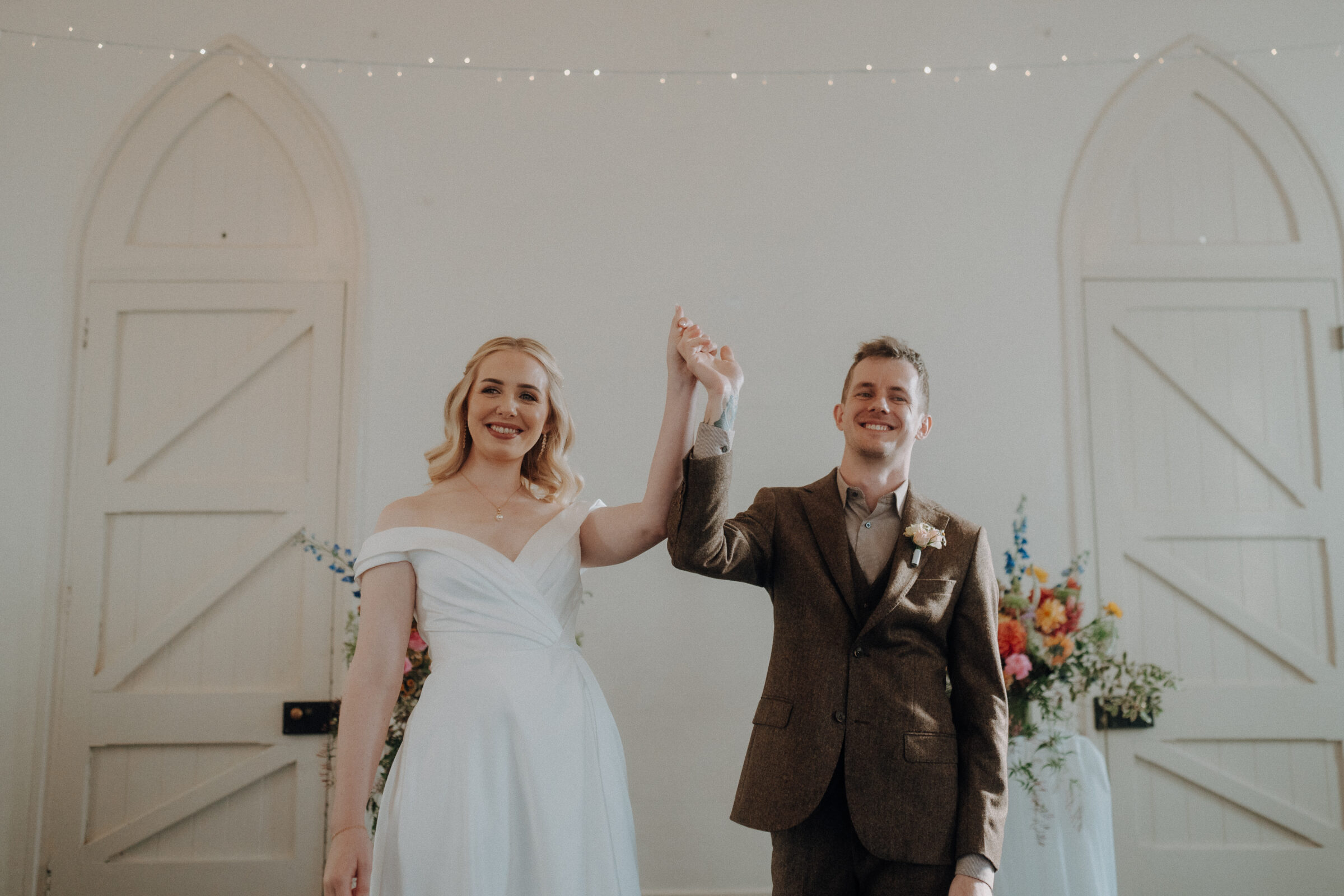 A couple celebrates their wedding, standing hand in hand. The bride wears a white dress, and the groom is in a brown suit. They stand in front of a light-colored background with floral arrangements.