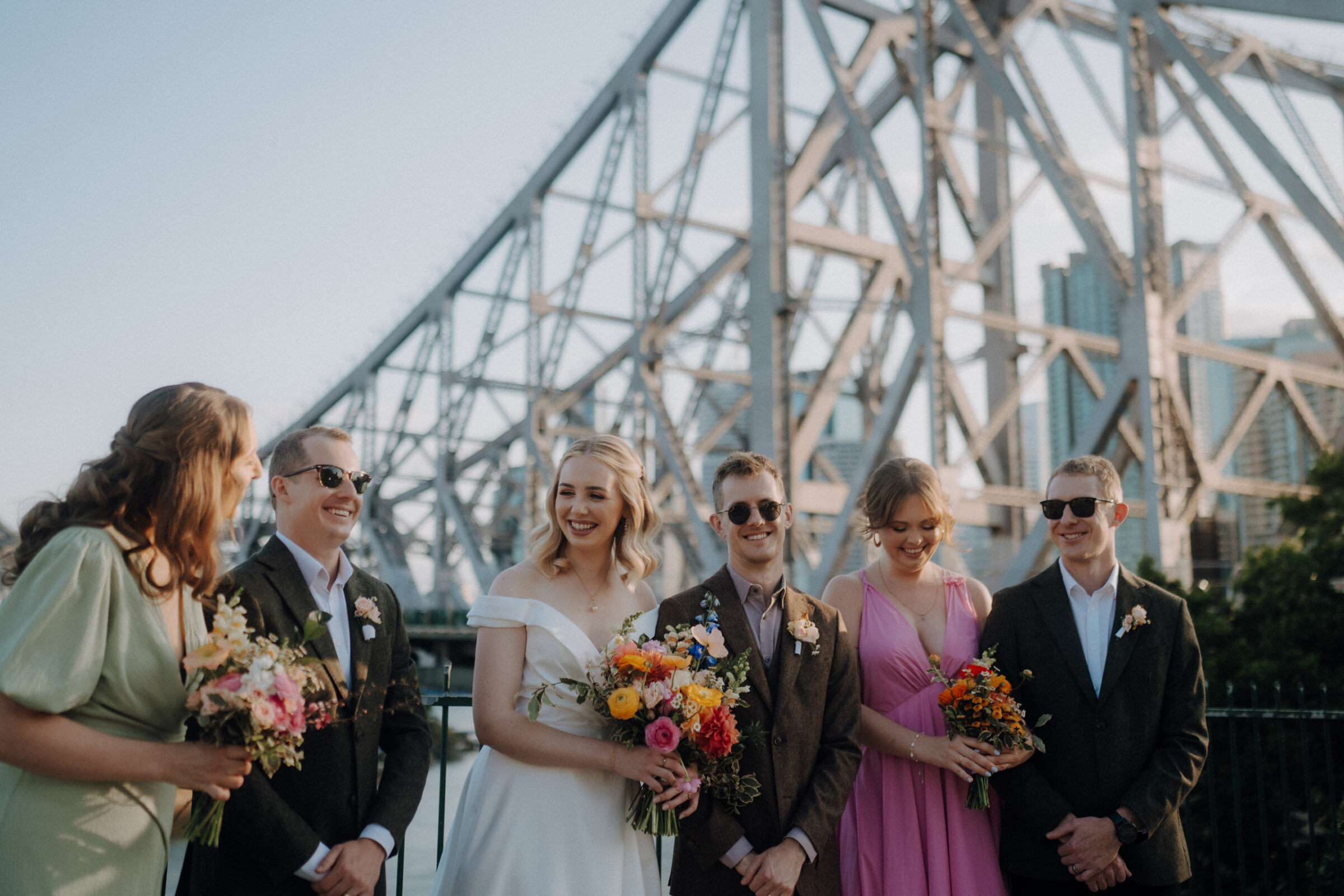 A group of people in formal attire stand together holding flowers, with a large metal bridge in the background.