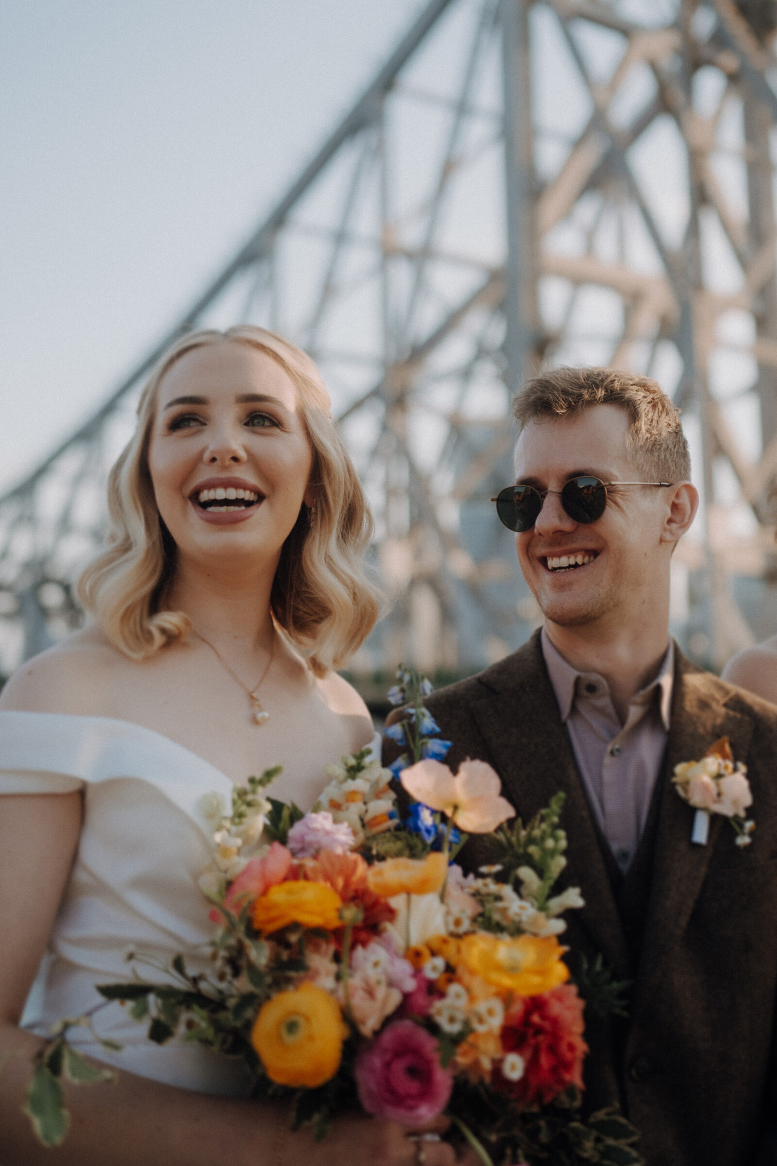 A smiling couple stands outdoors in front of a bridge. The woman holds a colorful bouquet, and both are dressed formally.