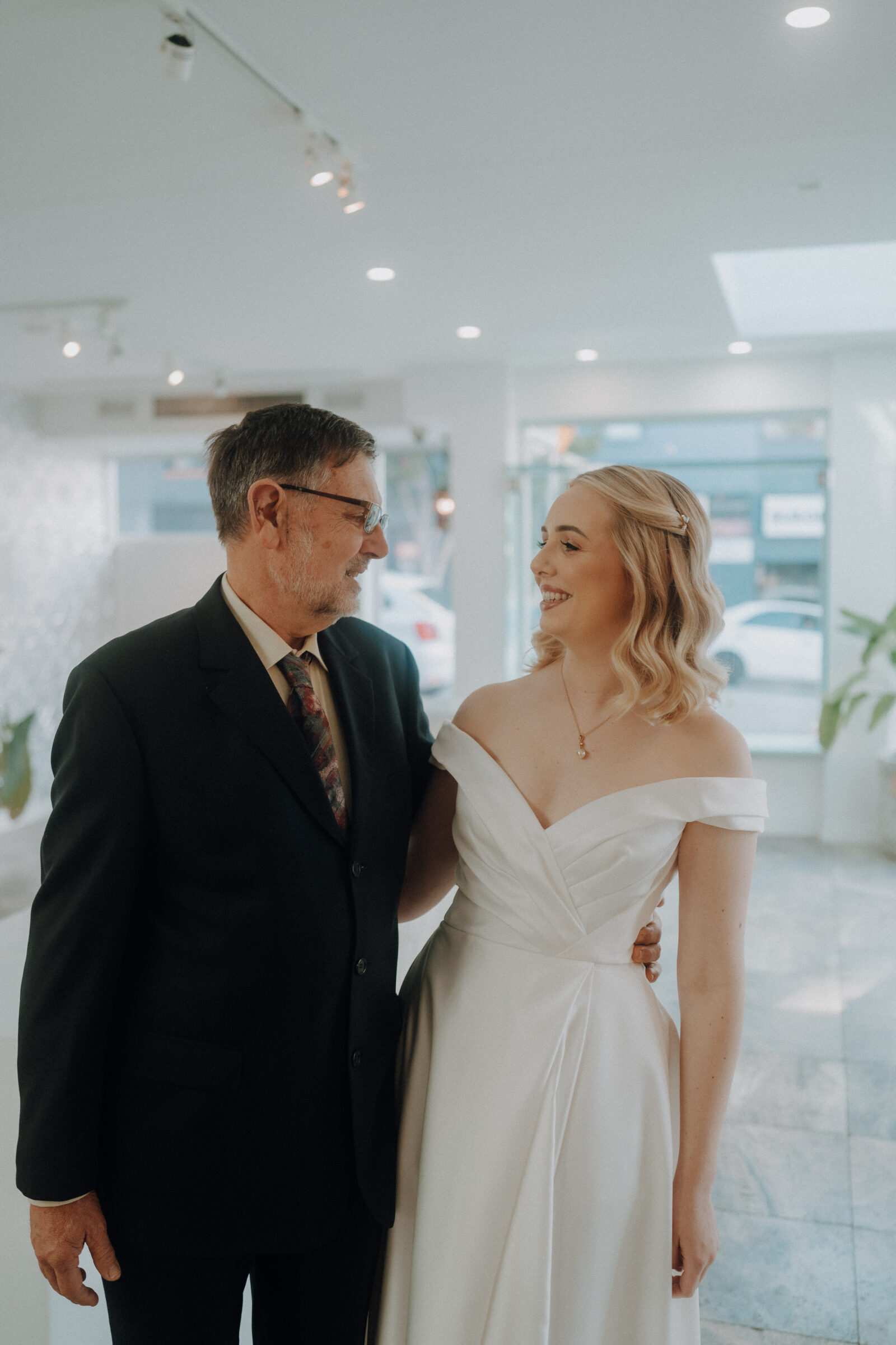 A man in a suit and a woman in a white off-the-shoulder dress stand indoors, smiling at each other.