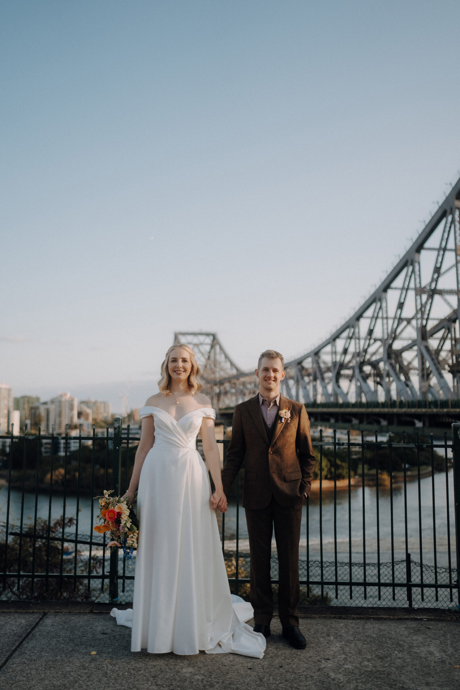 A couple stands holding hands, dressed in wedding attire, with a large steel bridge and cityscape in the background. The woman holds a colorful bouquet.