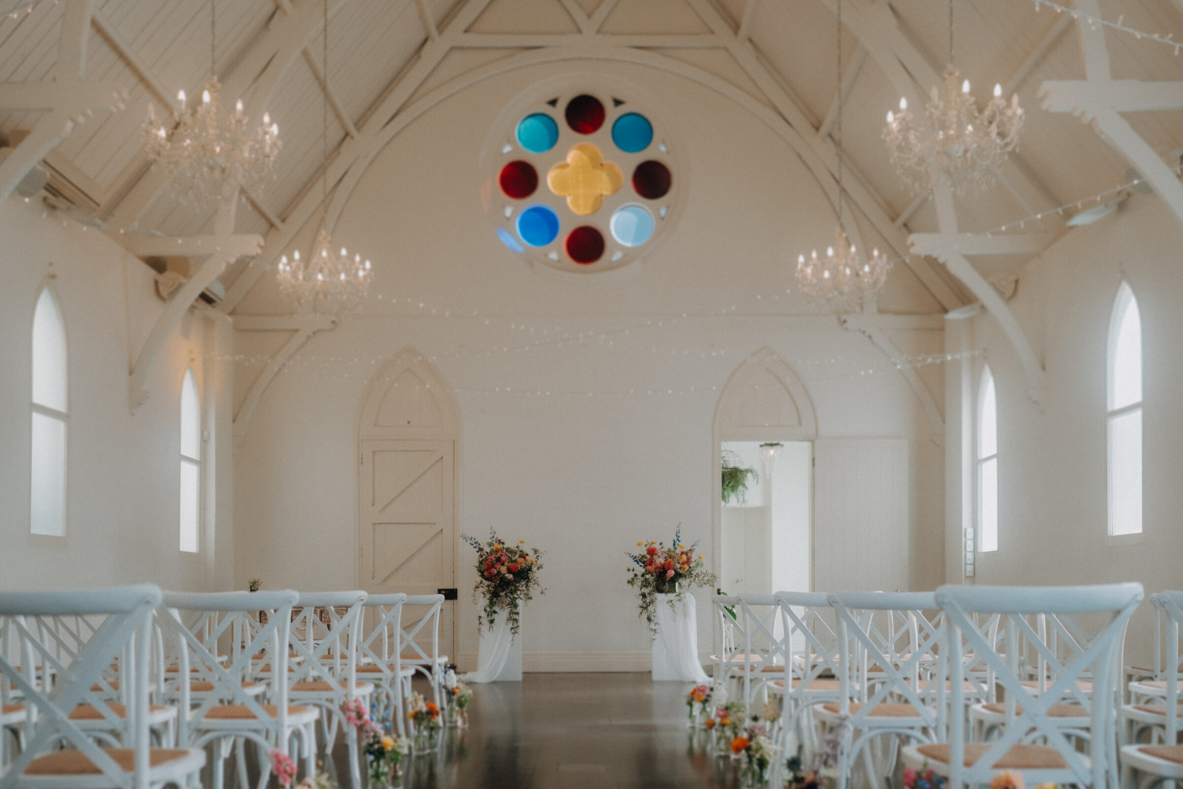 A white chapel interior with high ceilings, colorful stained glass window, floral arrangements, and rows of white chairs set up for a ceremony.