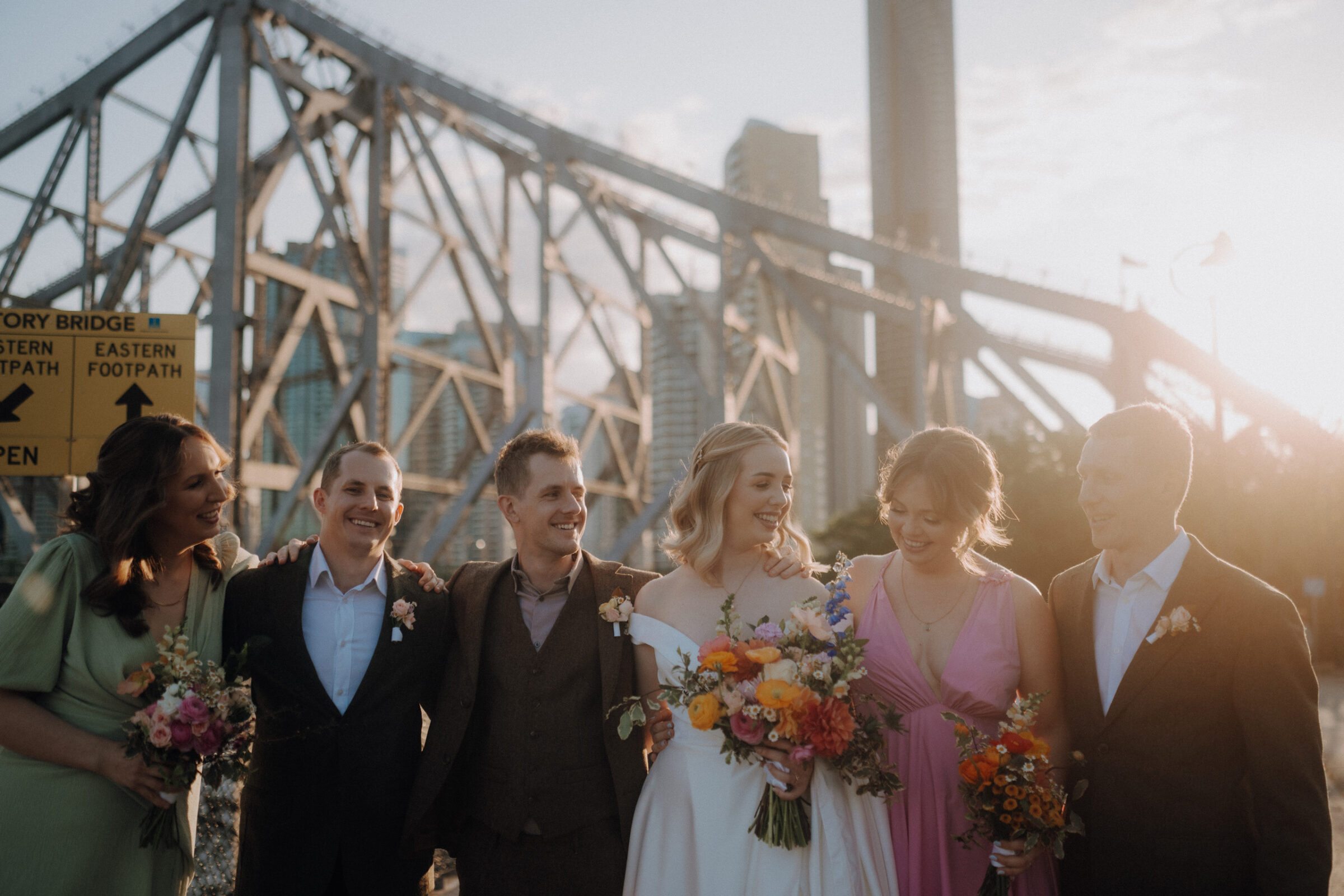 A group of six people in formal attire stands smiling in front of a large steel bridge, holding bouquets of colorful flowers.